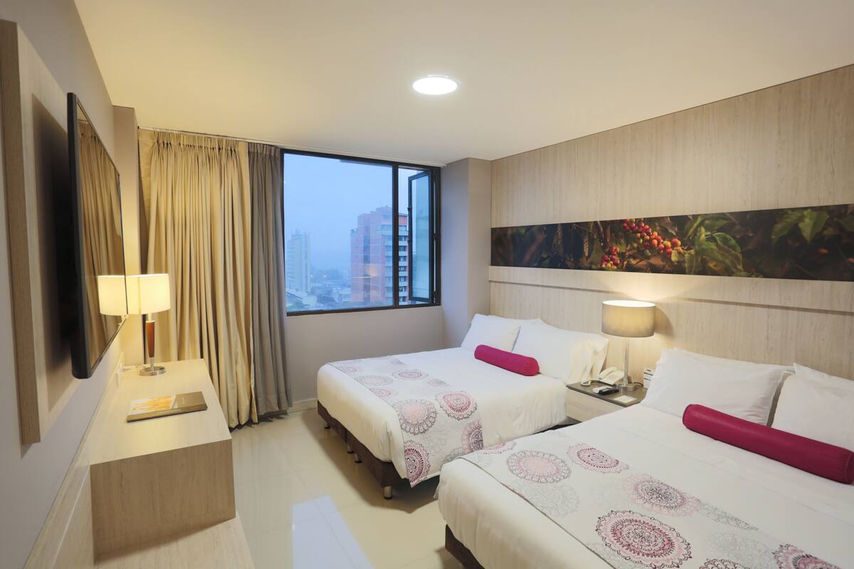 A hotel room with two beds, a desk, a chair, a window with curtains, a painting on the wall, and a city view.