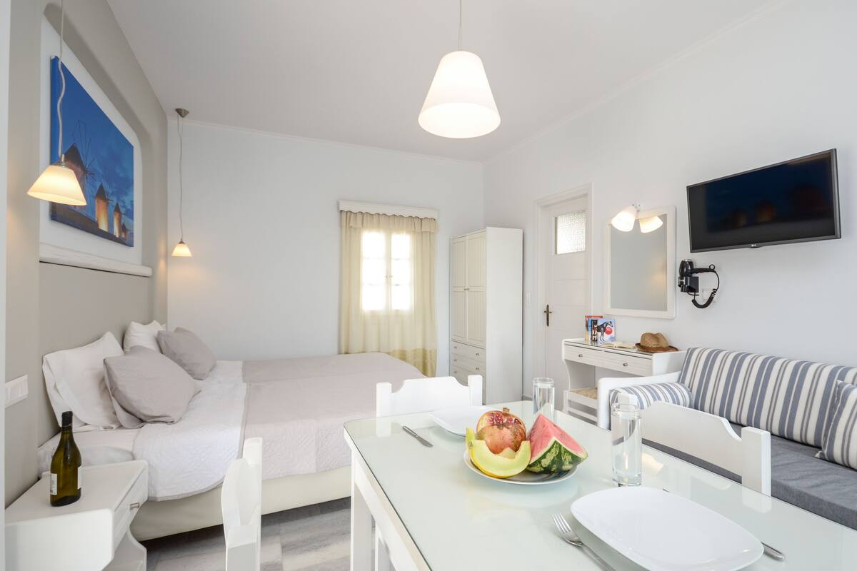 A modern hotel room with a bed, a dining table with breakfast, a sofa, and a TV.