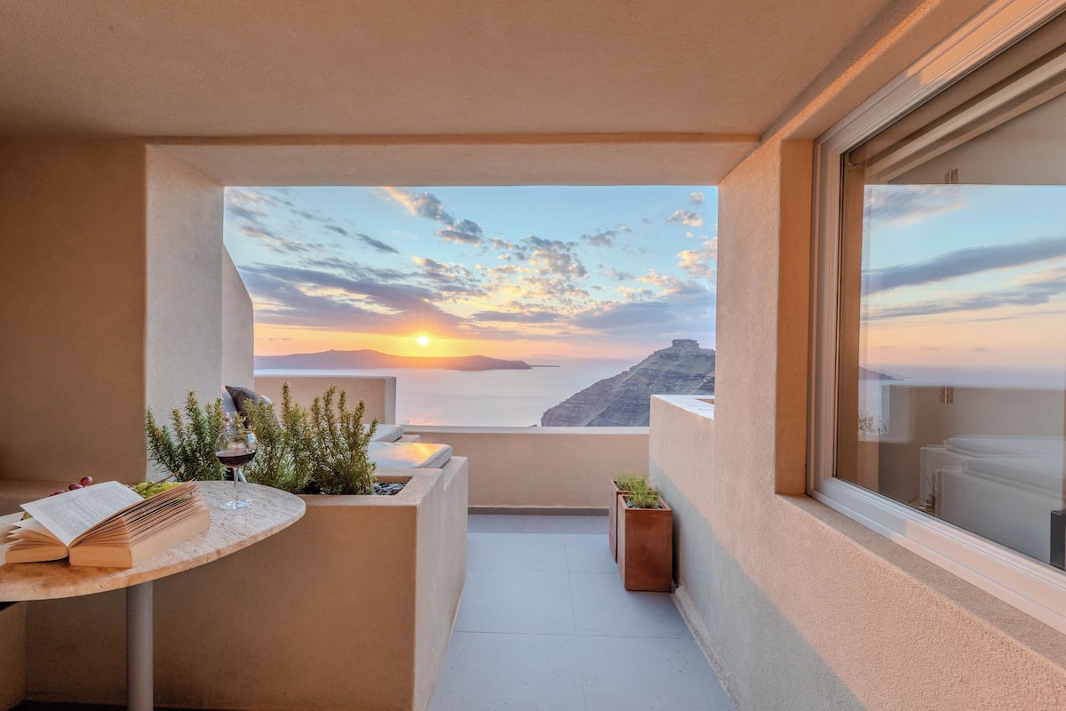 A balcony with a view of the sunset over the sea, a table with a book and a glass of wine, and potted plants.