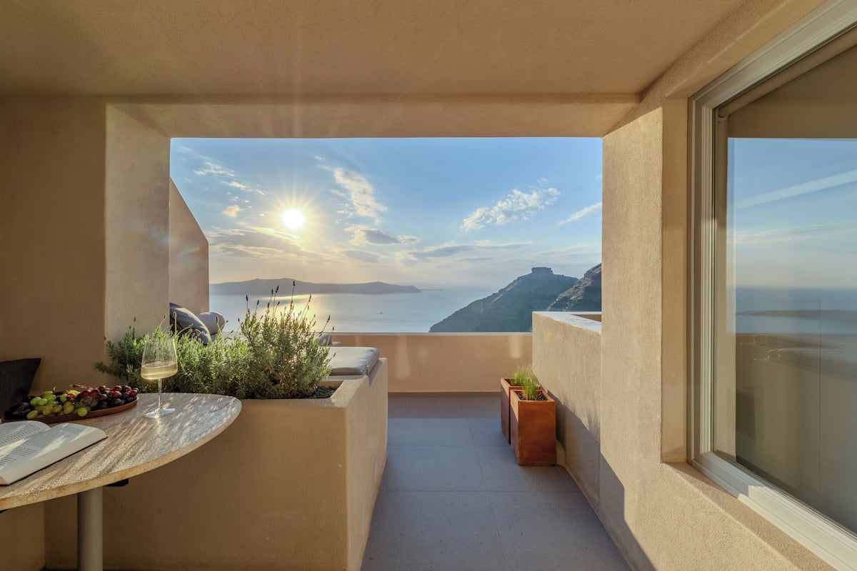 A balcony with a view of the sea and mountains, a table with a book and a glass of wine, and potted plants.