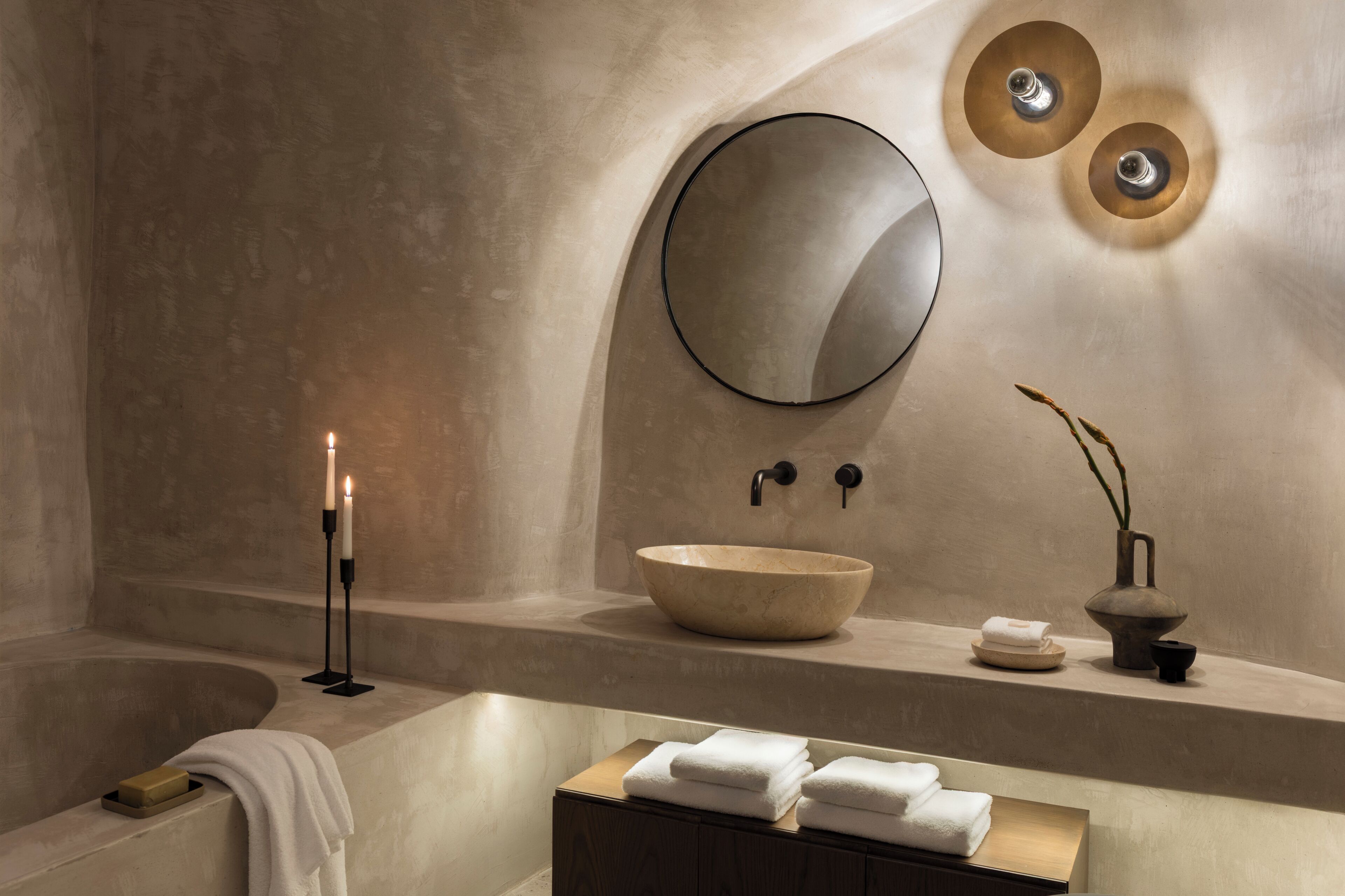 A modern bathroom with a large bathtub, a round mirror, a wooden bowl sink, and a candle on a stand.