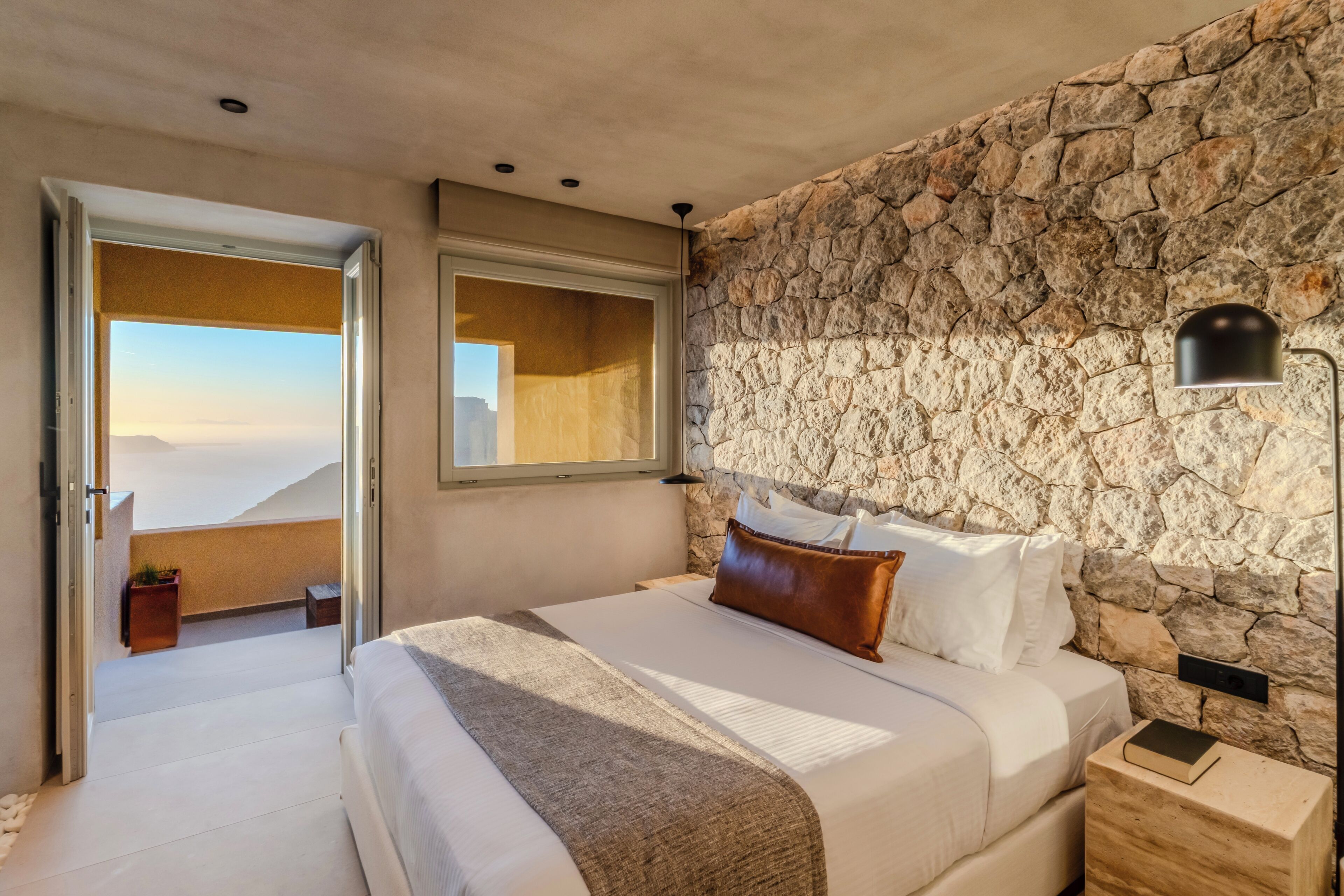 A bedroom with a stone wall, a bed with white linens, a nightstand, and a window with a view.