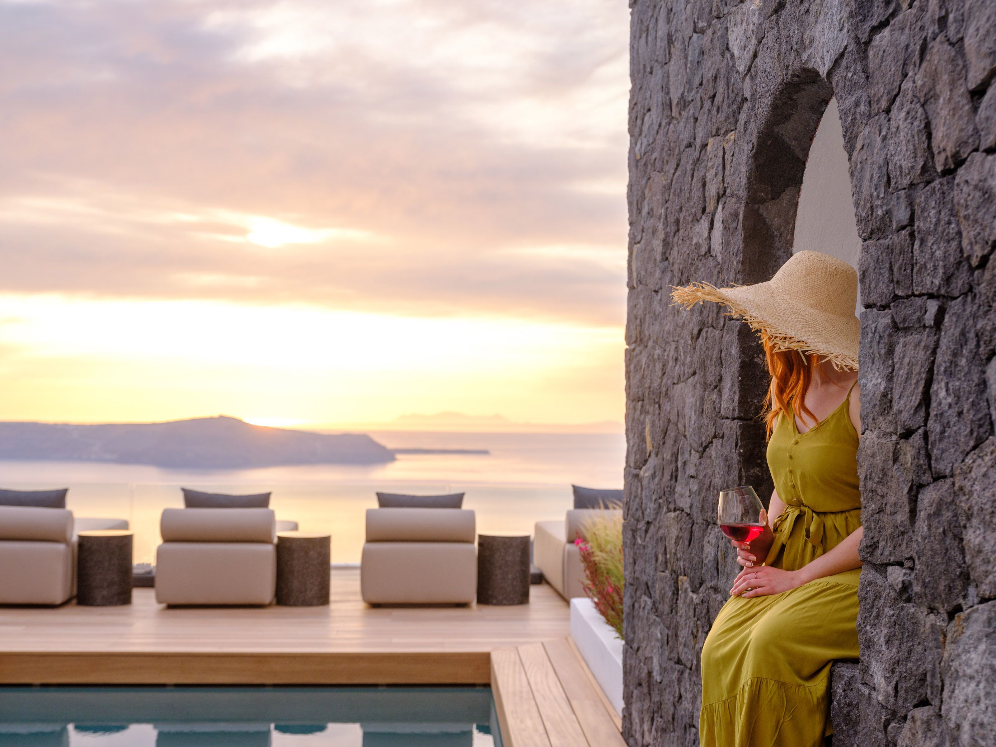 A woman in a yellow dress and straw hat sitting by a pool with a view of the sunset and mountains.