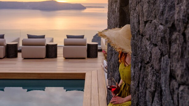 Una persona con un vestido verde sentada junto a una piscina con vista al atardecer, una pared de piedra y sillones de descanso.