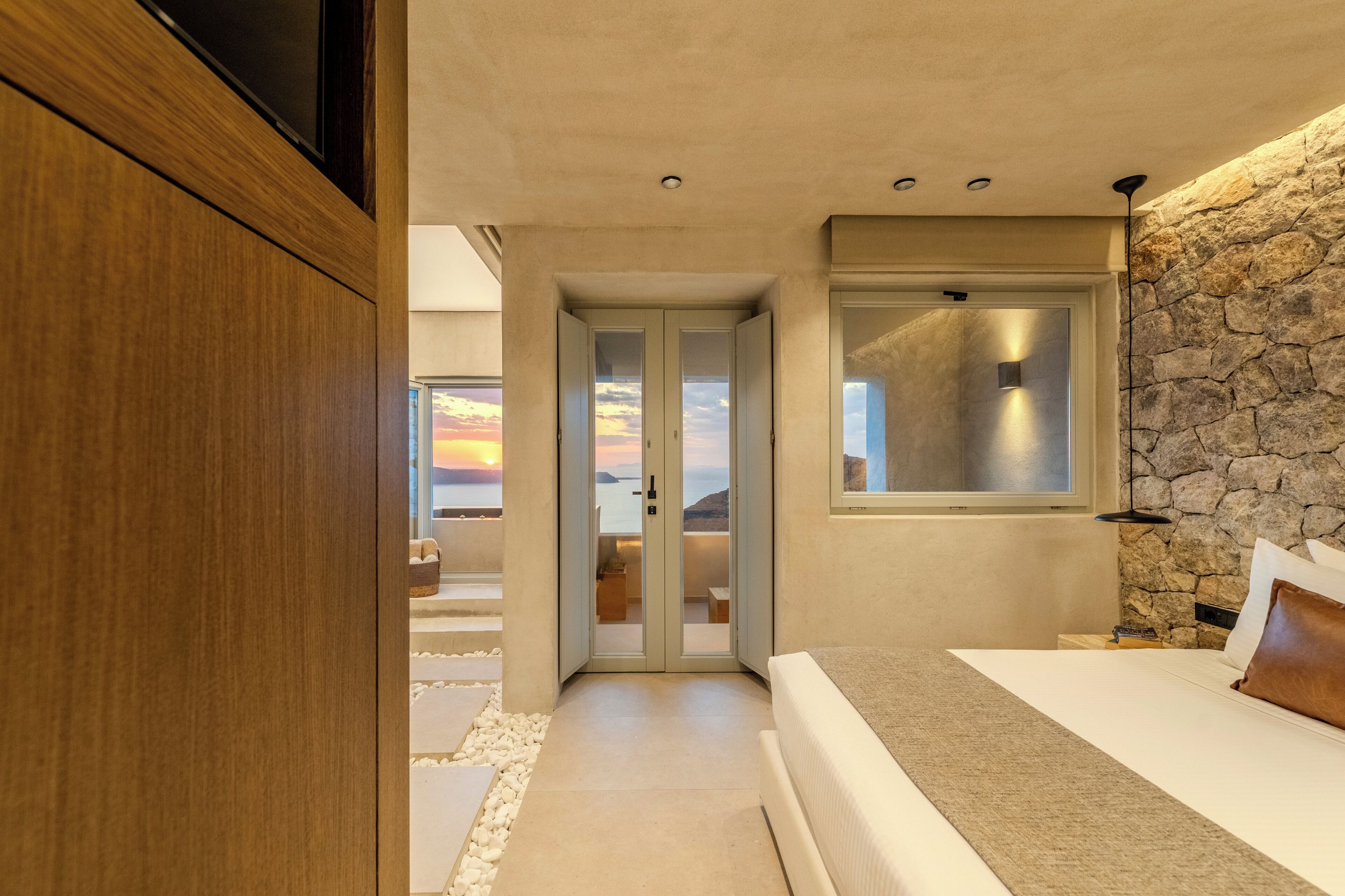 Un dormitorio moderno con una pared de piedra, una cama, una mesita de noche y vistas al atardecer.