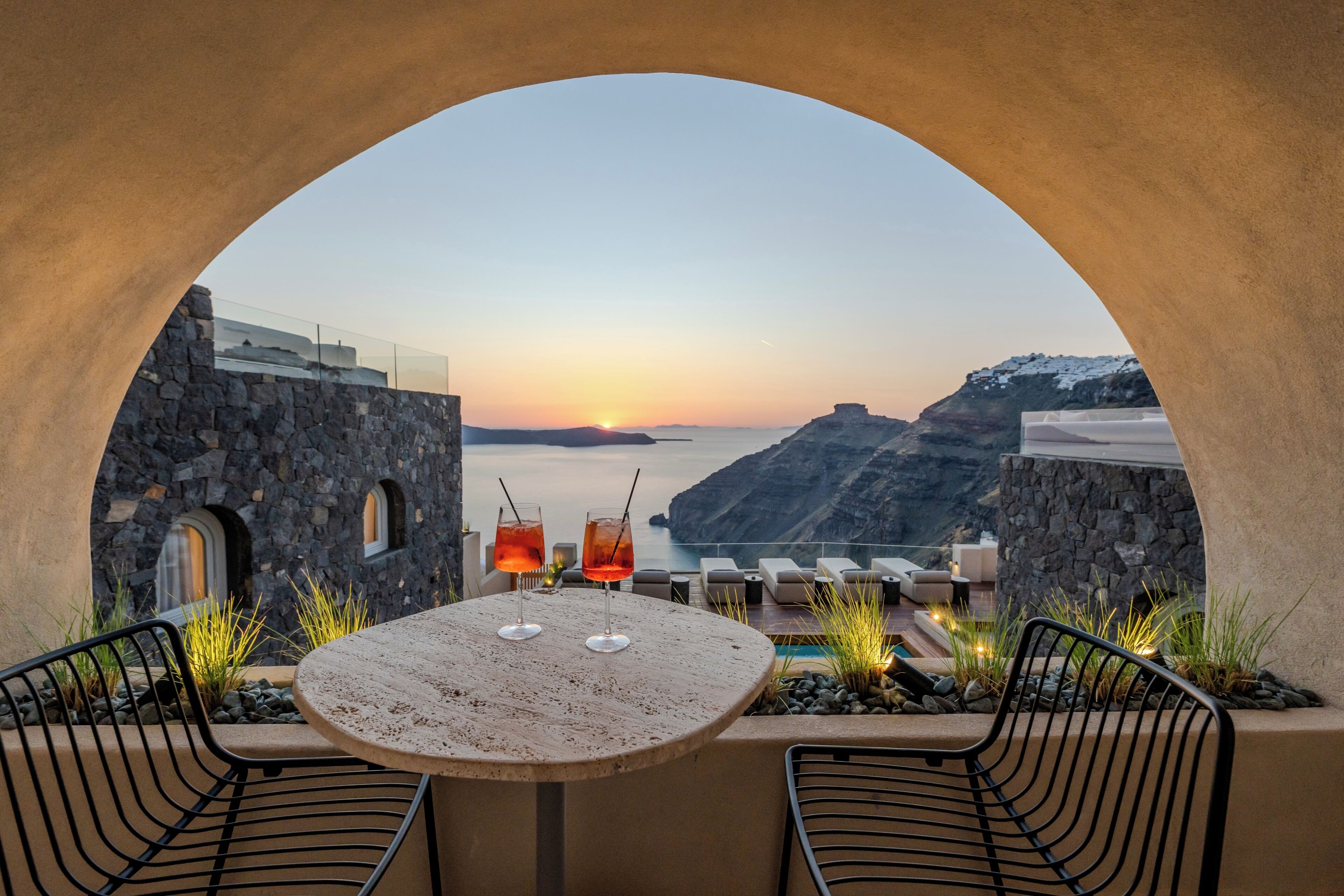 A table with two glasses, a stone wall, and a view of the sea at sunset.