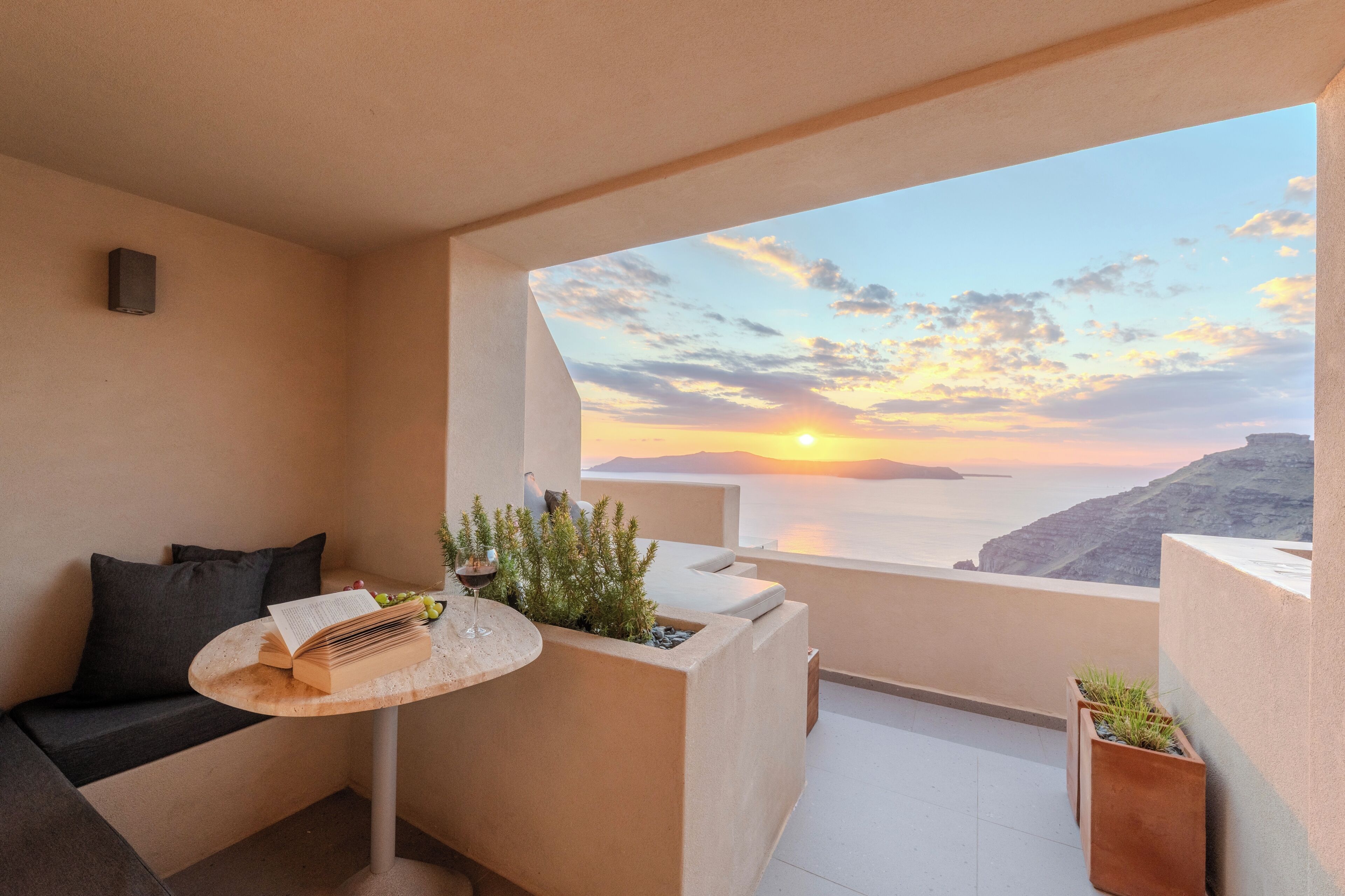A balcony with a view of the sunset, a sofa, a small table with a book and a glass of wine, and a potted plant.