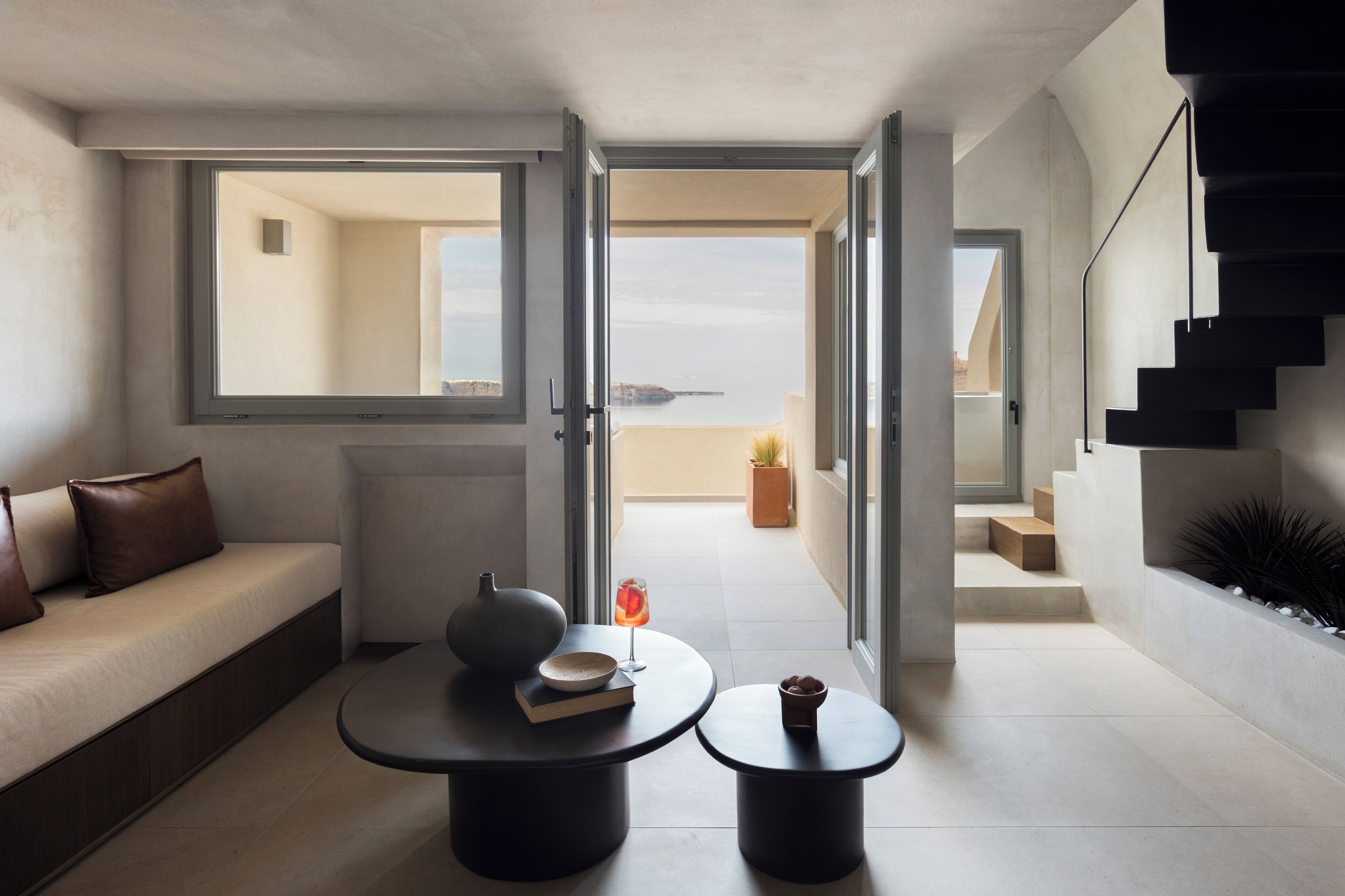 Un intérieur moderne avec un canapé, deux tables rondes, un escalier et une vue sur l’extérieur.