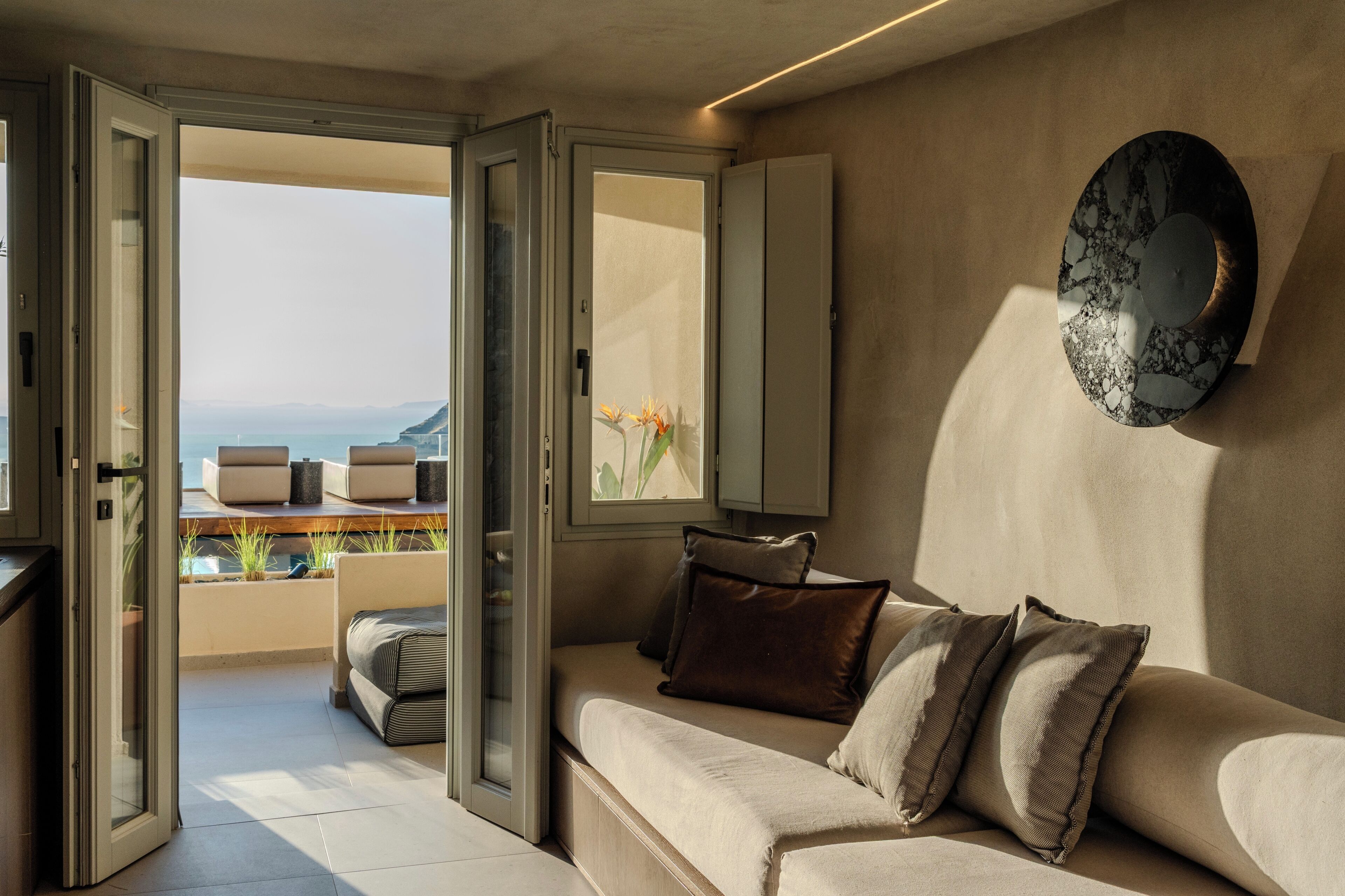 An open sliding door leading to a balcony with a view of the sea, a sofa with cushions, and a decorative plate on the wall.