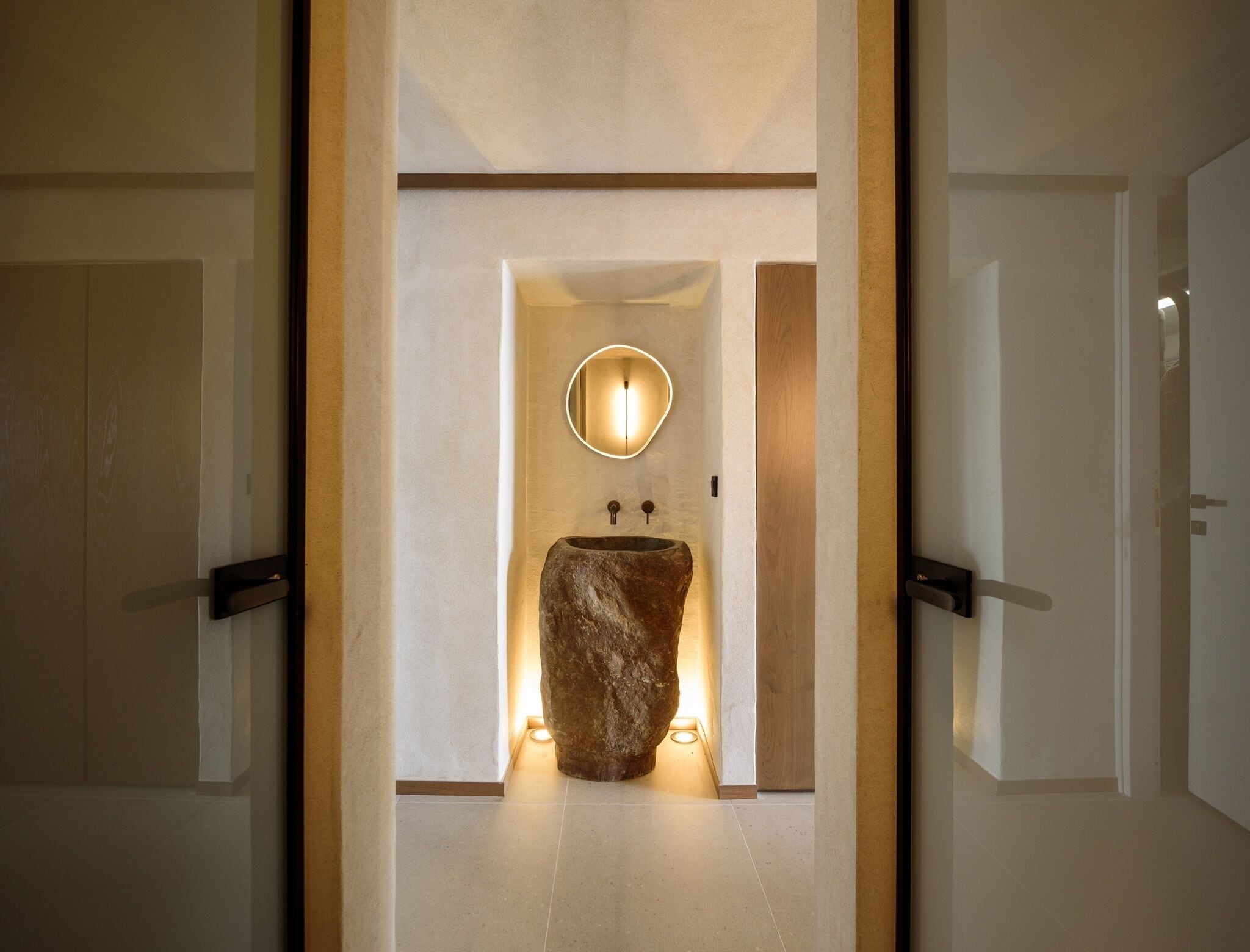 A modern bathroom with a large stone sink, a round mirror, and a wooden door frame.