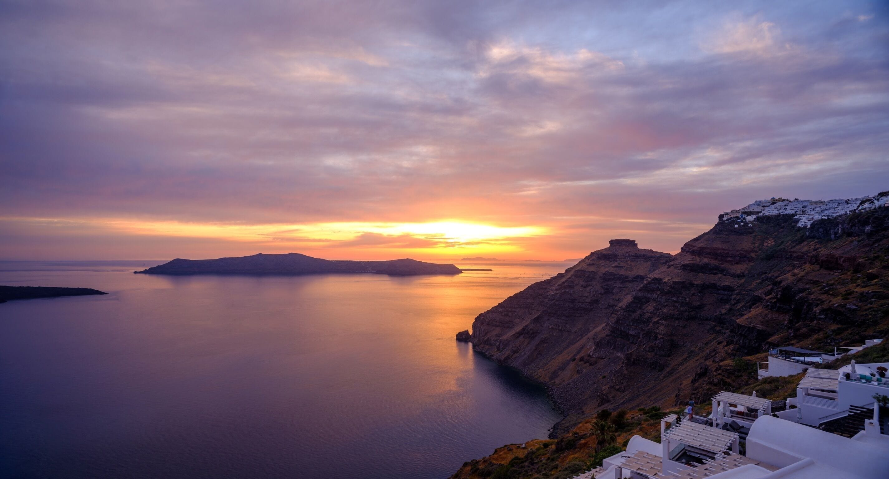 Un paisaje costero con un atardecer sobre el mar, que presenta edificios blancos con techos planos y un acantilado rocoso.