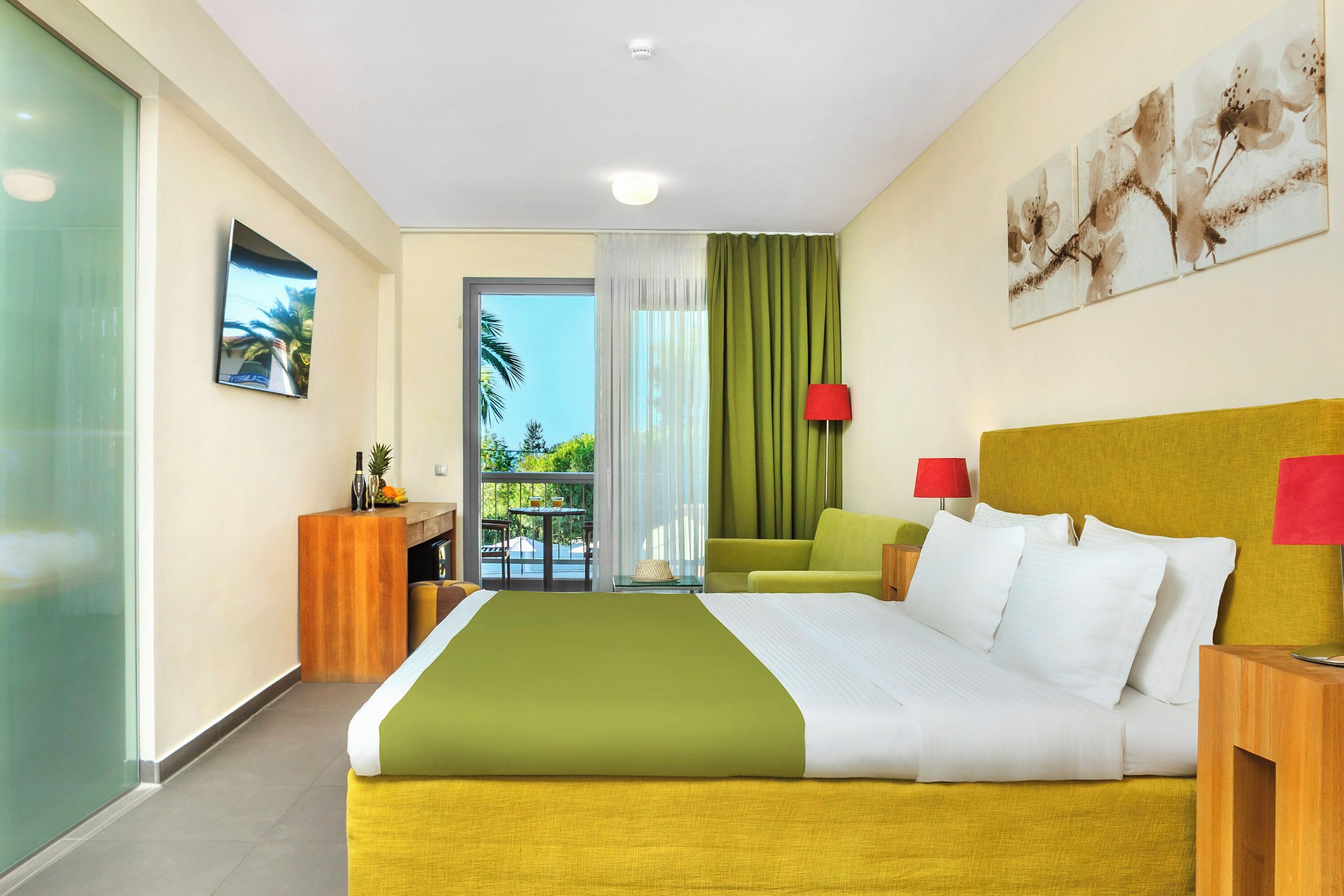 A hotel room with a bed, a desk, a chair, a TV, a balcony with a view of palm trees, and a painting on the wall.