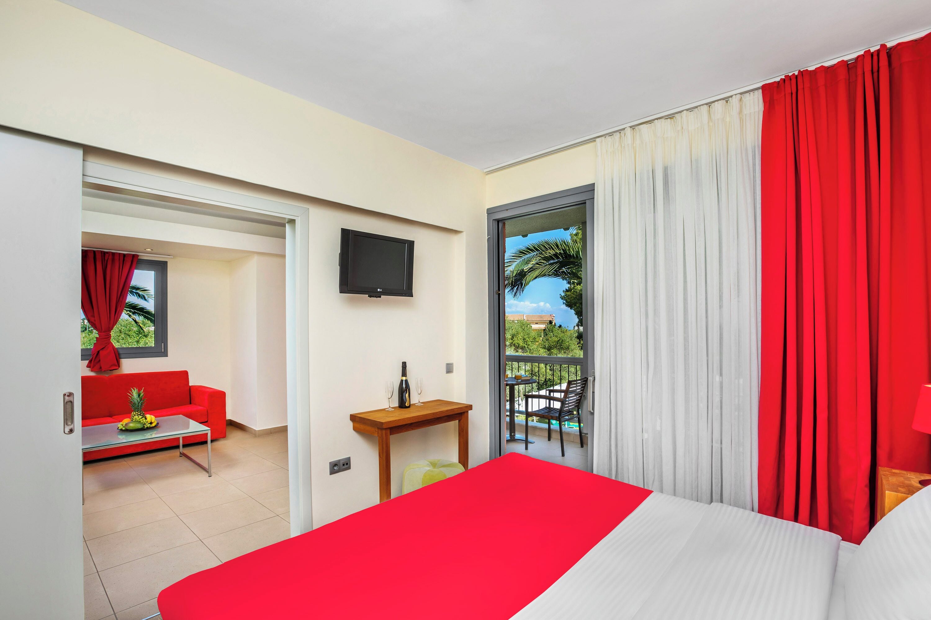 A hotel room with a bed, a red sofa, a small table with a bottle and glasses, a TV on the wall, and a balcony with a view of greenery.
