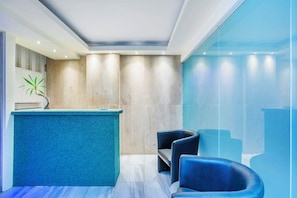 A reception area with a blue reception desk, seating area with chairs, and a large window with a view of the outside. - Hanioti Village Resort (Kassandra)