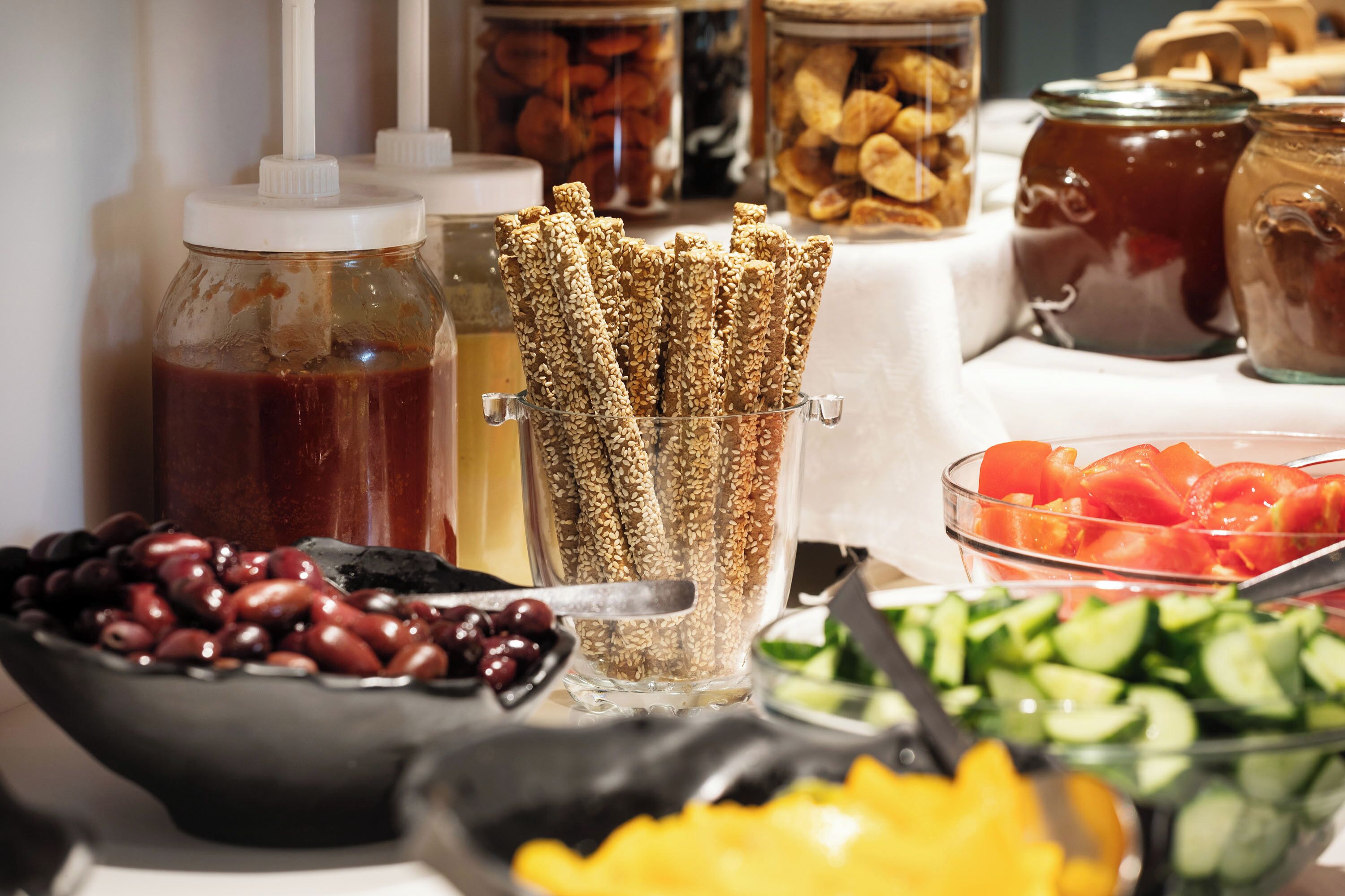 Une table de buffet proposant divers aliments, notamment des olives, des tomates, des concombres et un bocal de graines de sésame.