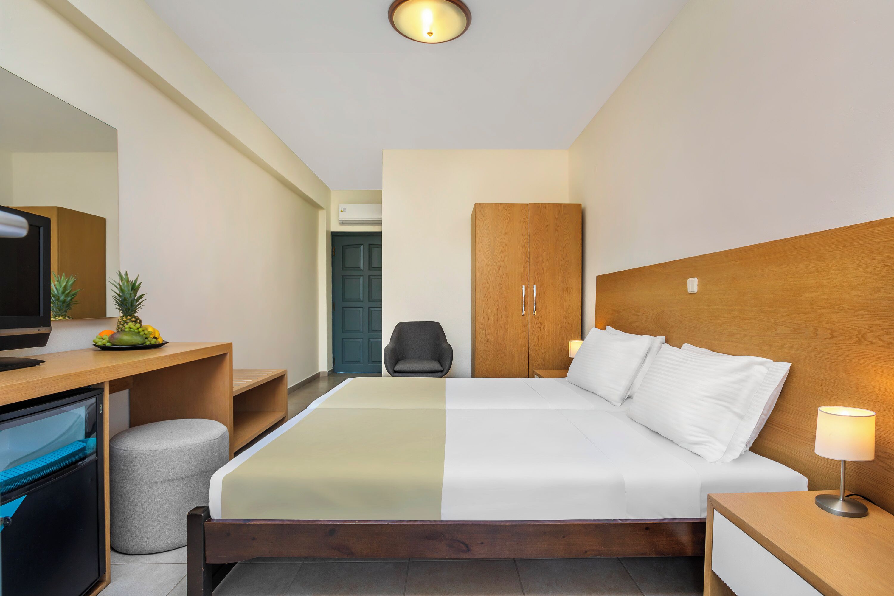 A hotel room with a bed, a desk with a television, a wardrobe, and a small stool.