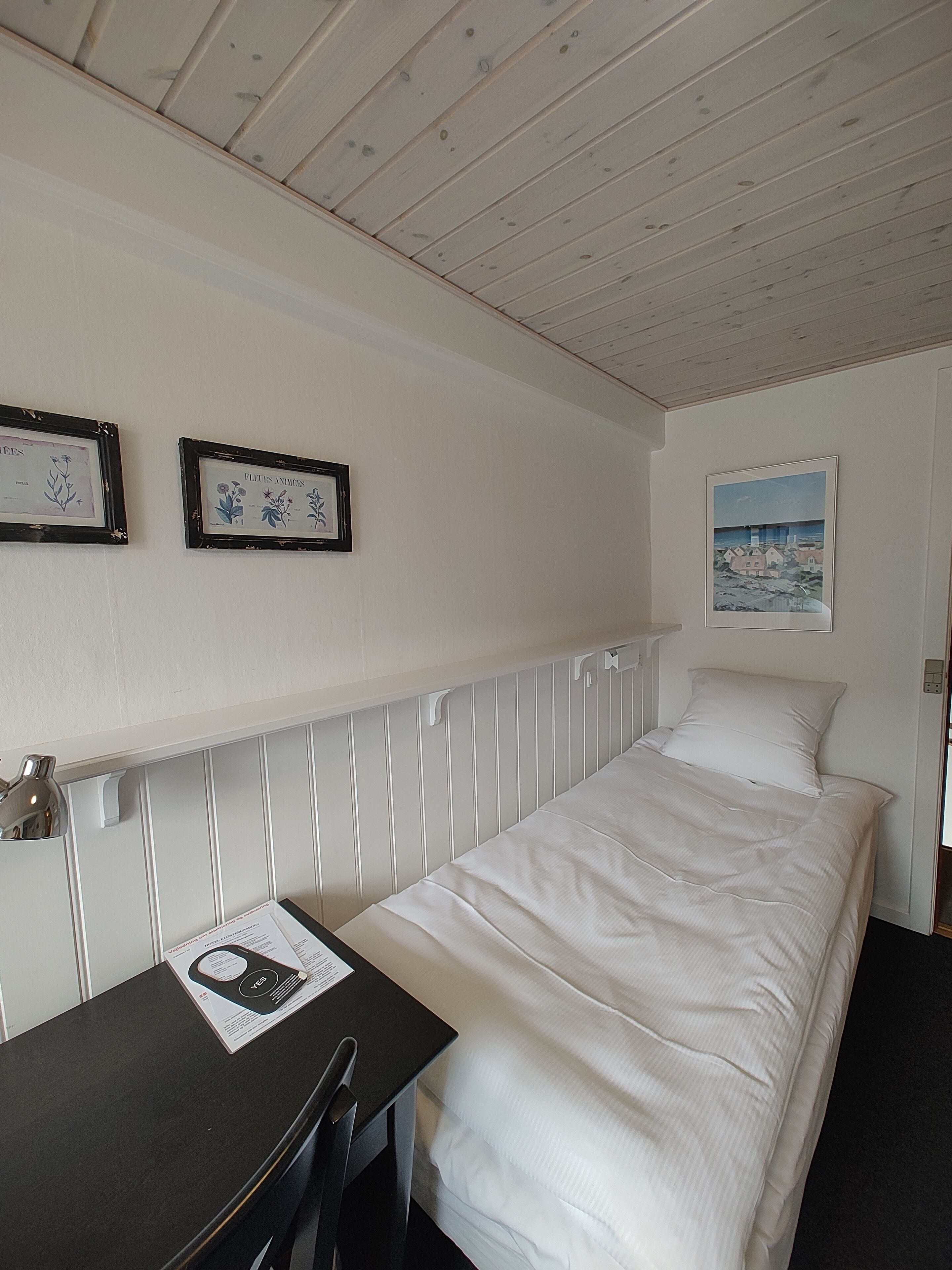 Una cama individual con ropa de cama blanca, una mesita con un menú, y dos láminas botánicas enmarcadas en la pared.