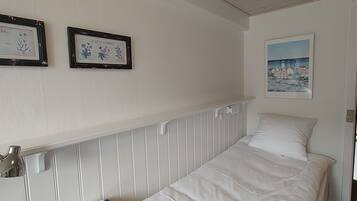 Ein Einzelbett mit weißer Bettwäsche, ein kleiner Tisch mit einer Speisekarte und zwei gerahmte Pflanzenbilder an der Wand.