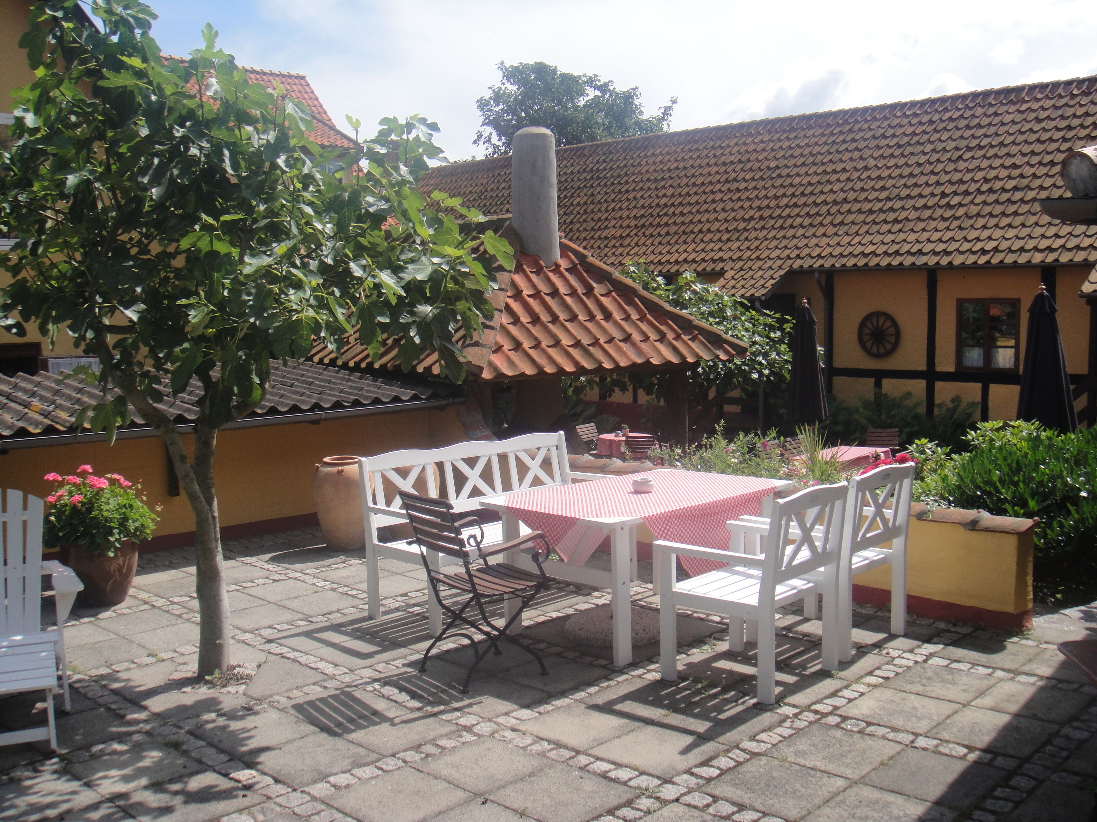 An outdoor patio with a table and chairs, surrounded by potted plants and a building with a tiled roof.
