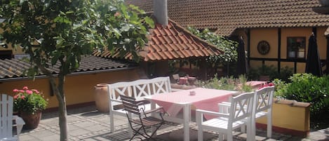 An outdoor patio with a table and chairs, surrounded by potted plants and a building with a tiled roof.