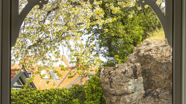 A view through a window with a decorative frame, featuring a garden with a rock formation, a hedge, and two lawn chairs. - Hotel Klostergaarden (Allinge)
