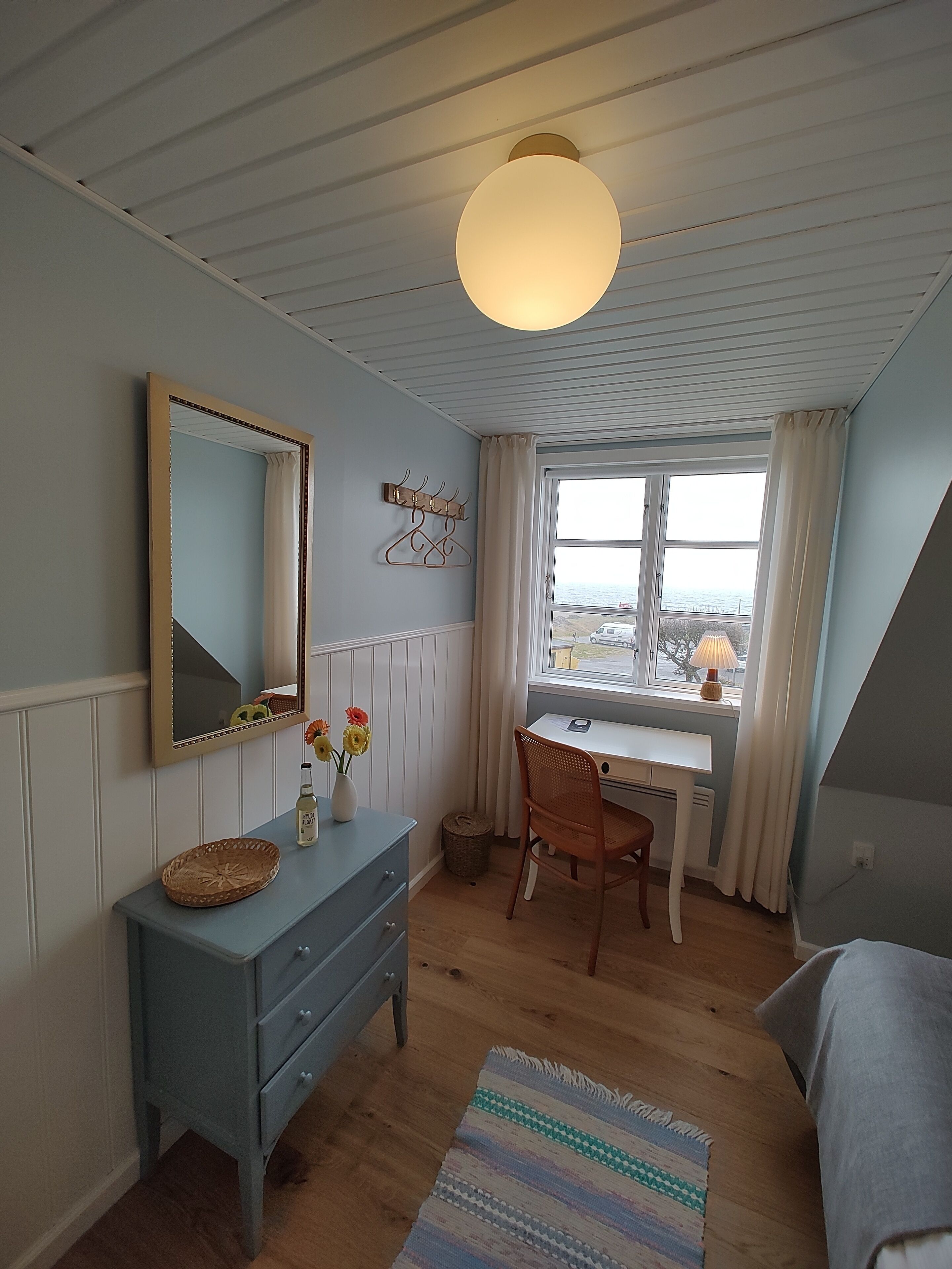 Une chambre à coucher avec une commode en bois, un bureau, une chaise, un grand miroir et une fenêtre donnant sur la mer.