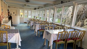 A dining area with multiple tables and chairs, large windows, and a ceiling fan. - Hotel Klostergaarden (Allinge)