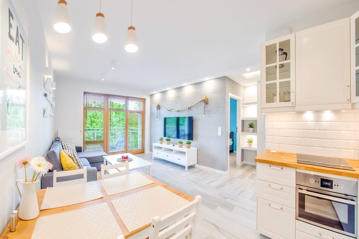 A modern kitchen with white cabinets, a wooden countertop, and built-in appliances. There is a dining area with a table and chairs, and a living room with a sofa and a TV.