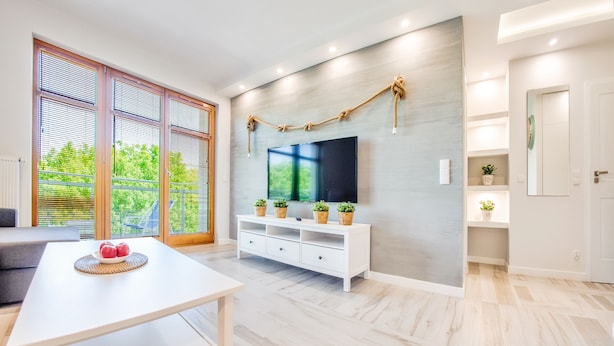 A modern living room with a white TV stand, a dining table with a fruit bowl, and large windows with blinds. - Apartments Sopot (Sopot)