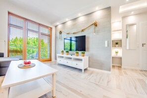 Una sala moderna con un mueble blanco para televisión, una mesa de comedor con frutero y ventanas grandes con persianas.