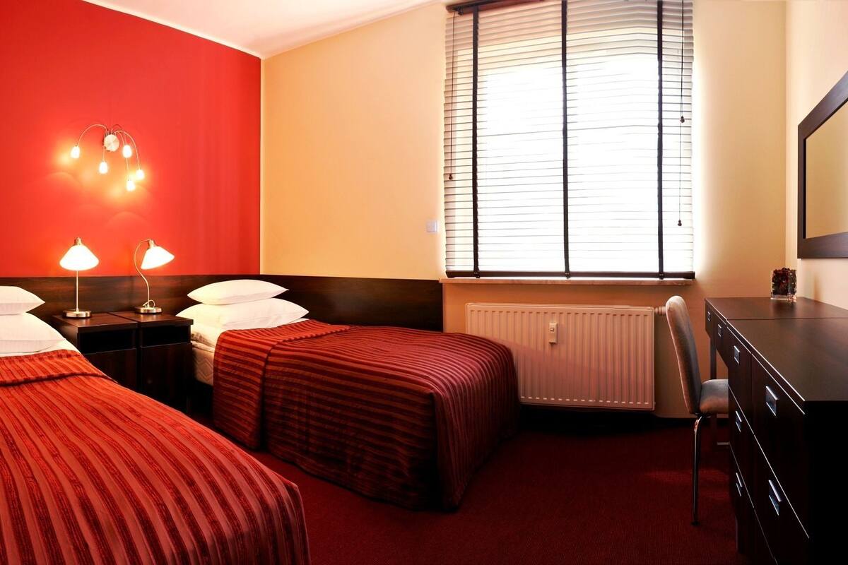 A hotel room with two single beds, a desk, a chair, and a window with blinds.
