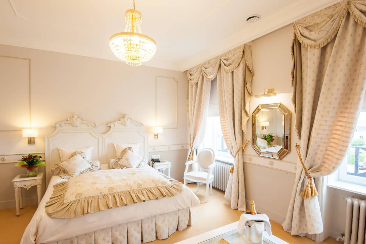 Una habitación de hotel con una cama grande, una araña, un espejo, un radiador y una ventana con cortinas.