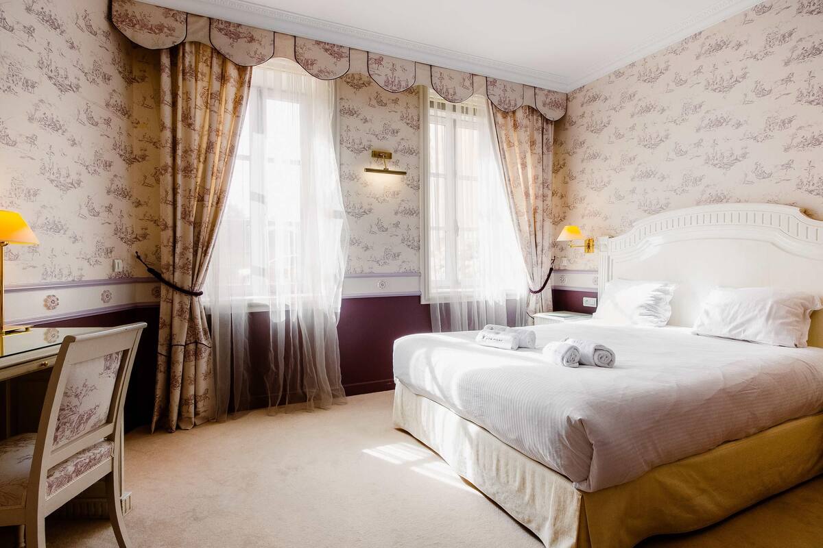 Une chambre d’hôtel avec un lit, un bureau et deux fenêtres équipées de rideaux.