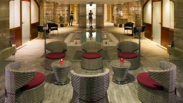 Un moderno vestíbulo de hotel con una piscina de cristal central, mobiliario de mimbre con cojines rojos y paredes de piedra.