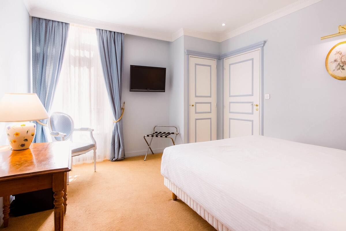 Une chambre d’hôtel avec un lit, un bureau avec une lampe, une chaise, un téléviseur fixé au mur et une fenêtre avec des rideaux.