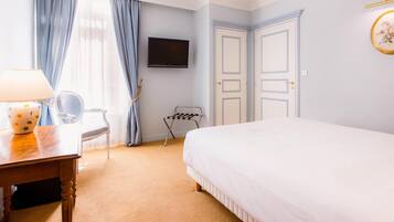 Habitación de hotel con cama, escritorio con lámpara, silla, televisor montado en la pared y ventana con cortinas.