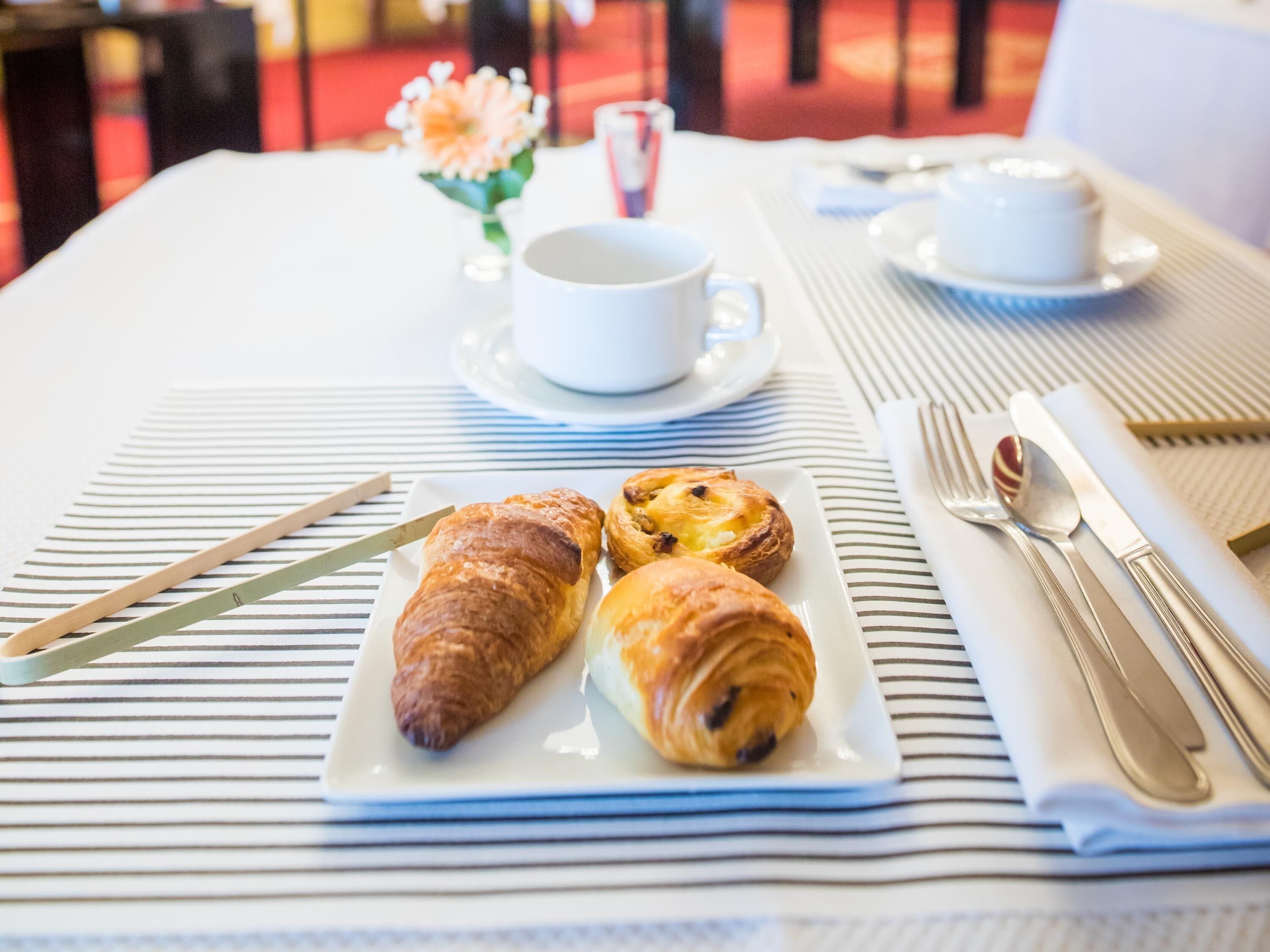 A breakfast setting with croissants on a plate, a cup of coffee, and a small vase with flowers.