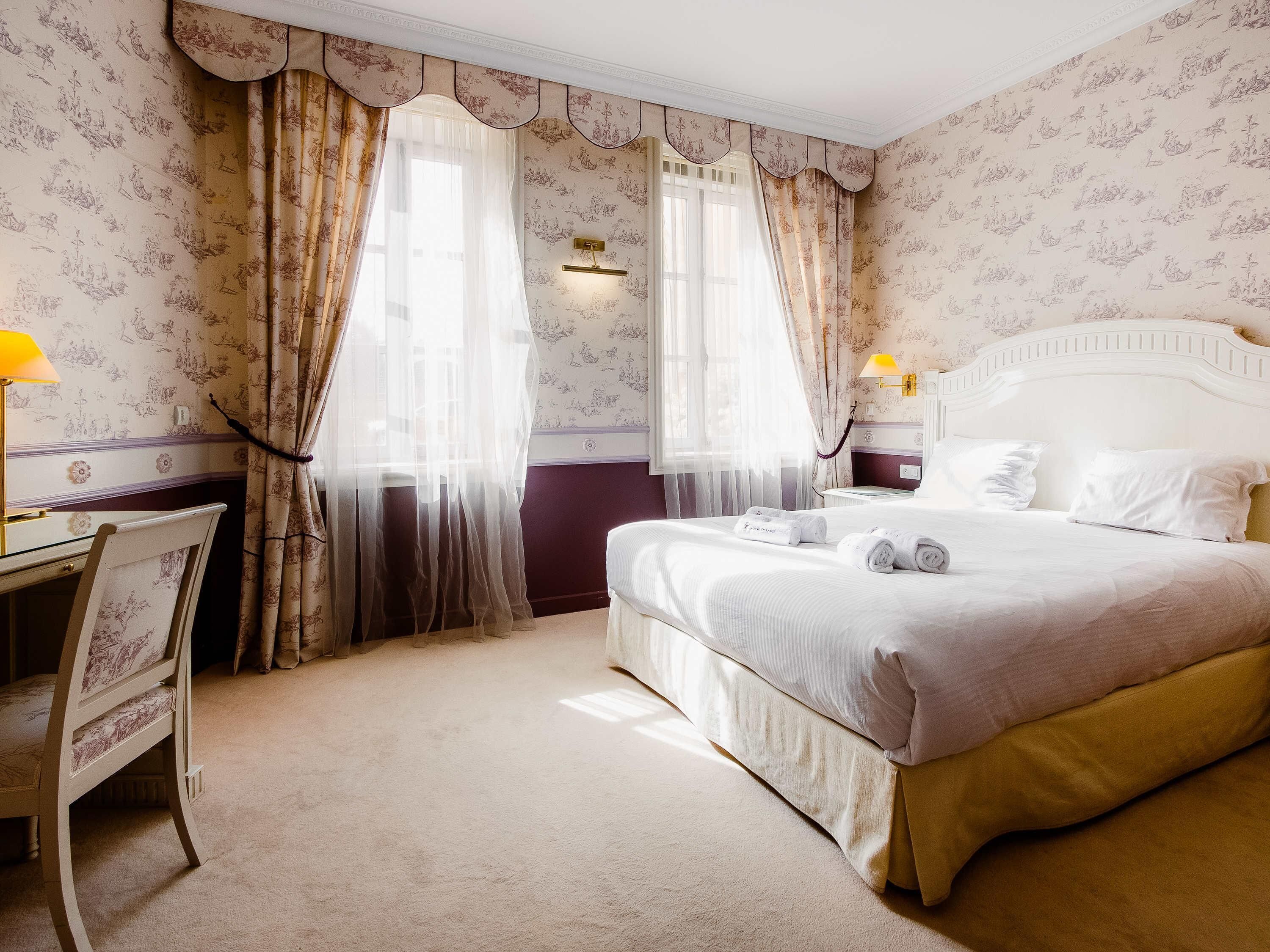 A hotel room with a bed, a desk, a chair, and two windows with curtains.
