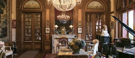 A grand room with a large chandelier, a grand piano, and a fireplace with a marble mantle.