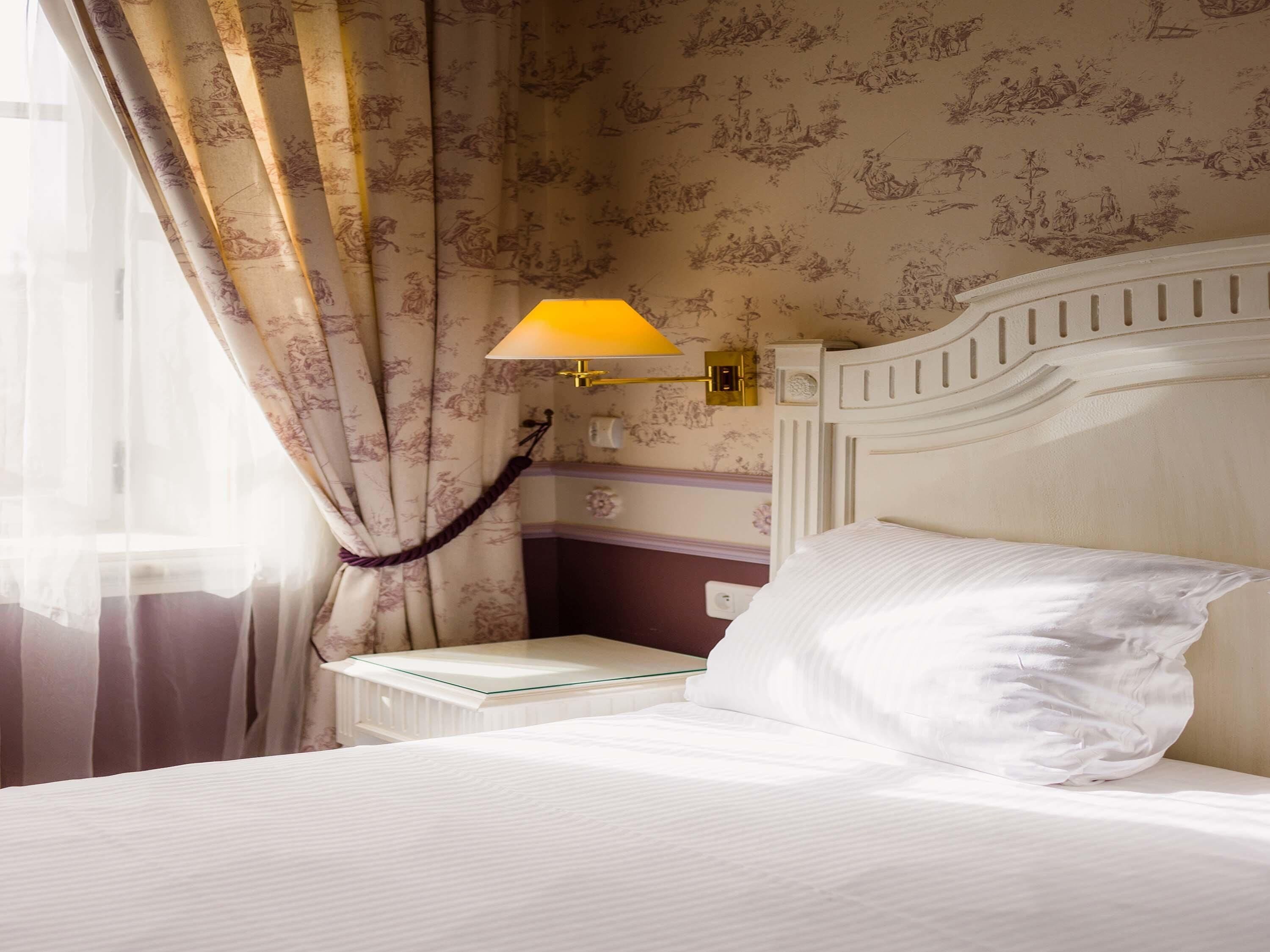 A neatly made bed with white linens, a white nightstand with a glass top, a wall-mounted lamp, and a patterned wallpaper.