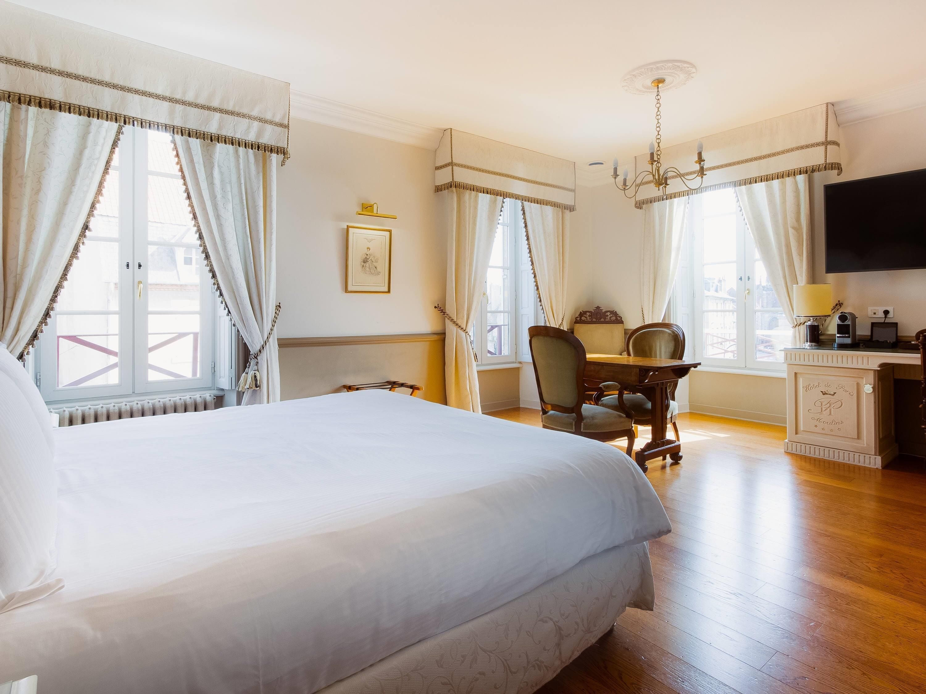 A hotel room with a large bed, a desk with a chair, a television, and a chandelier.