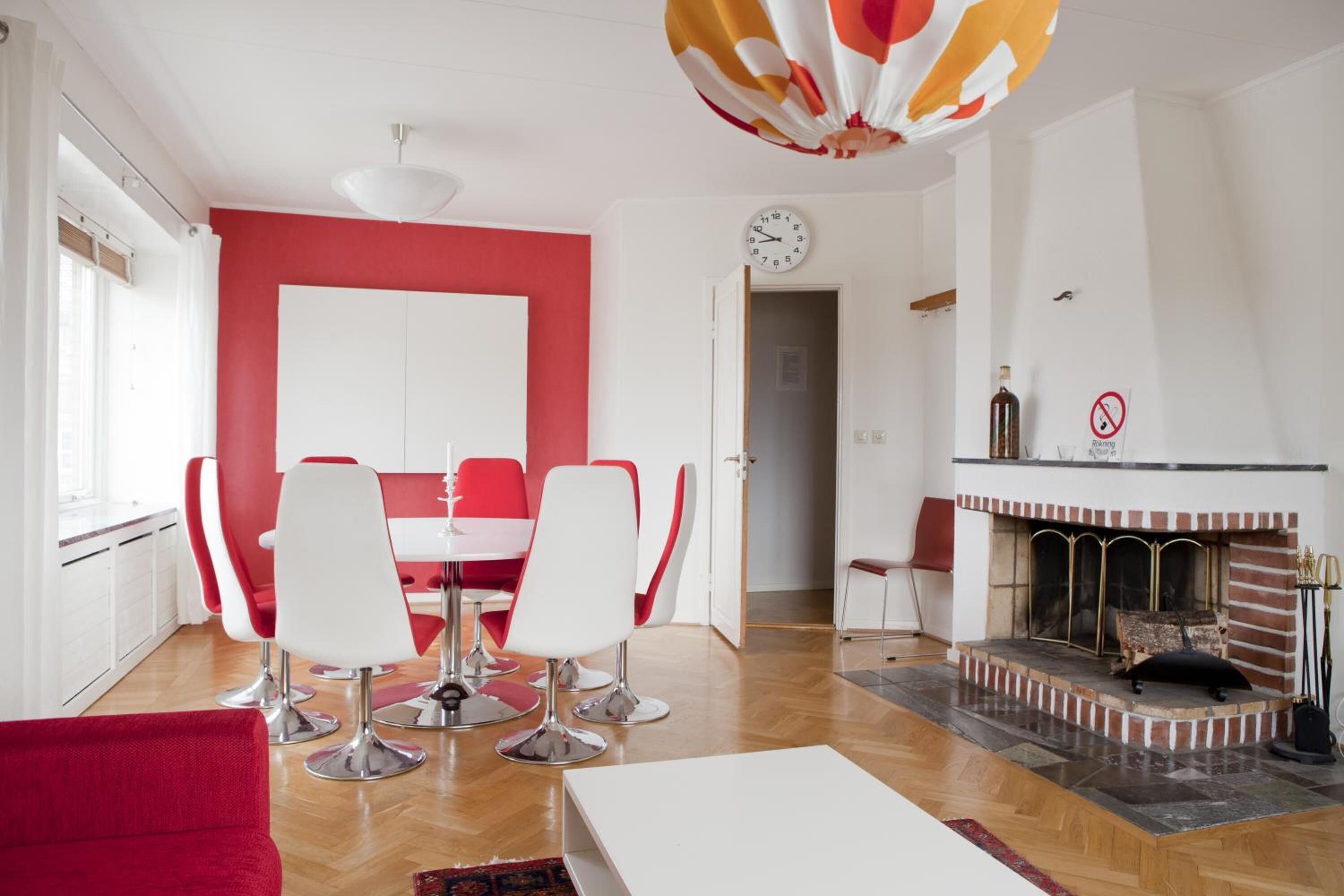 A modern living room with a red accent wall, a dining area with white chairs, a fireplace, and a clock on the wall.