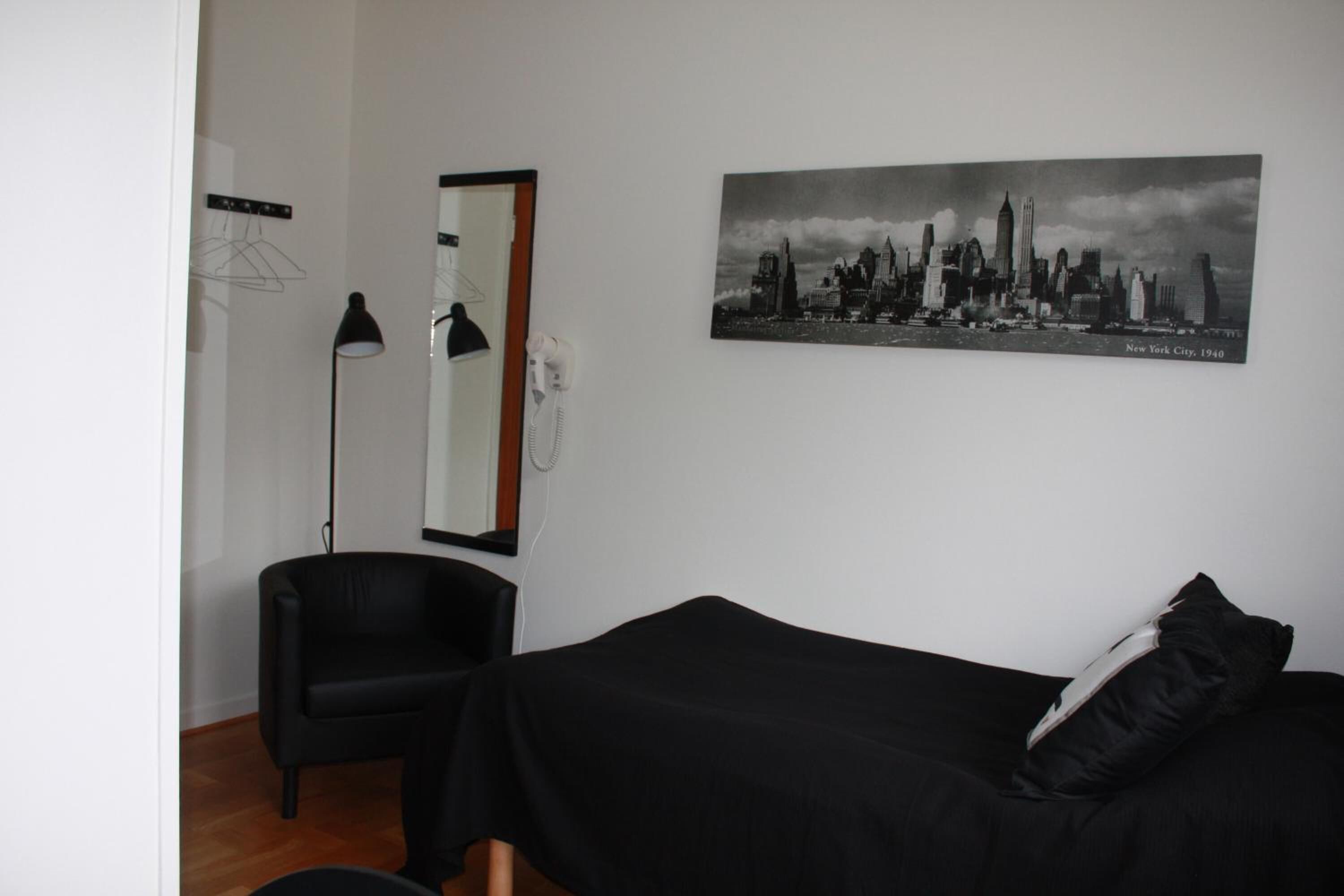 Ett rum med en säng, en stol, en lampa, en spegel och en tavla med en stadsbild på väggen.