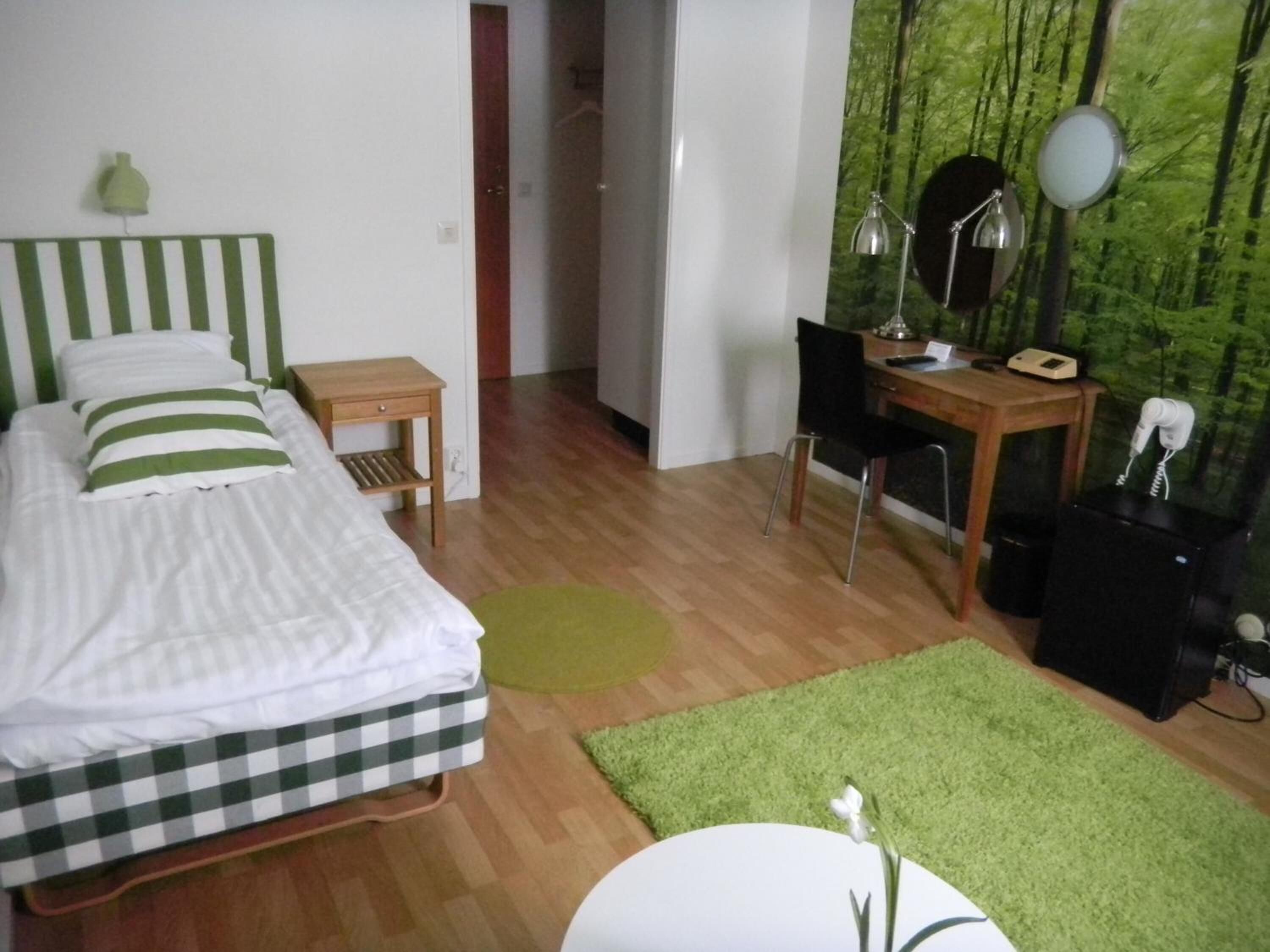 A bedroom with a bed, a desk, a chair, a small table, a lamp, and a wall-mounted mirror.