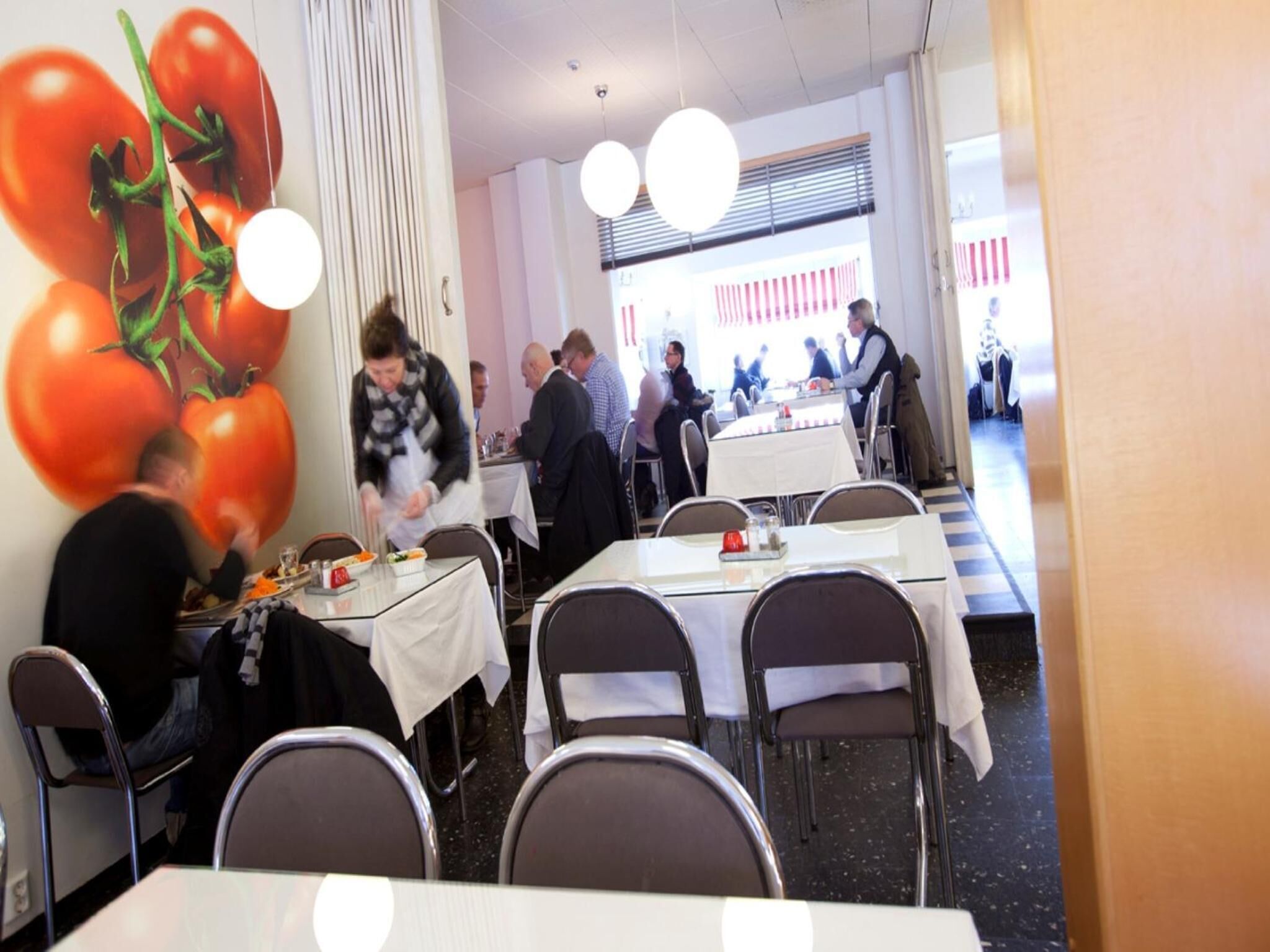 A restaurant with tables set for dining, a waiter serving food, and a large tomato mural on the wall.