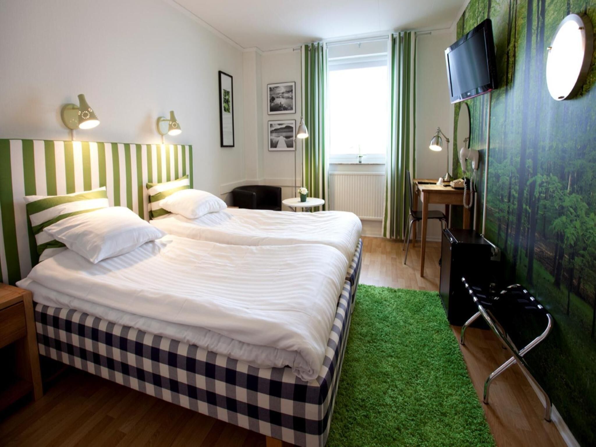 A hotel room with two beds, a desk, a chair, a television, and a green wall with a forest mural.