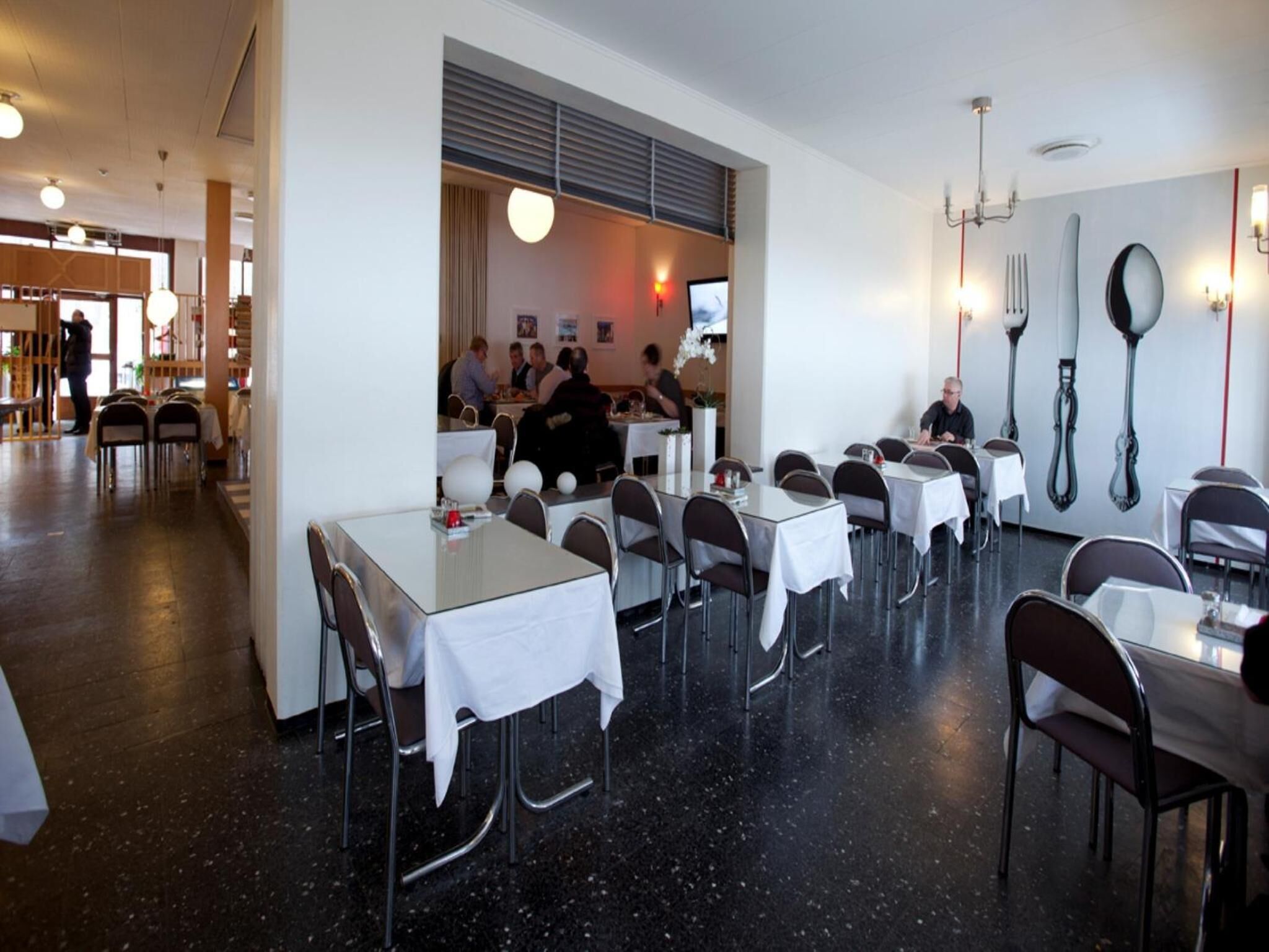A restaurant with tables and chairs, a counter with people, and a wall with cutlery.
