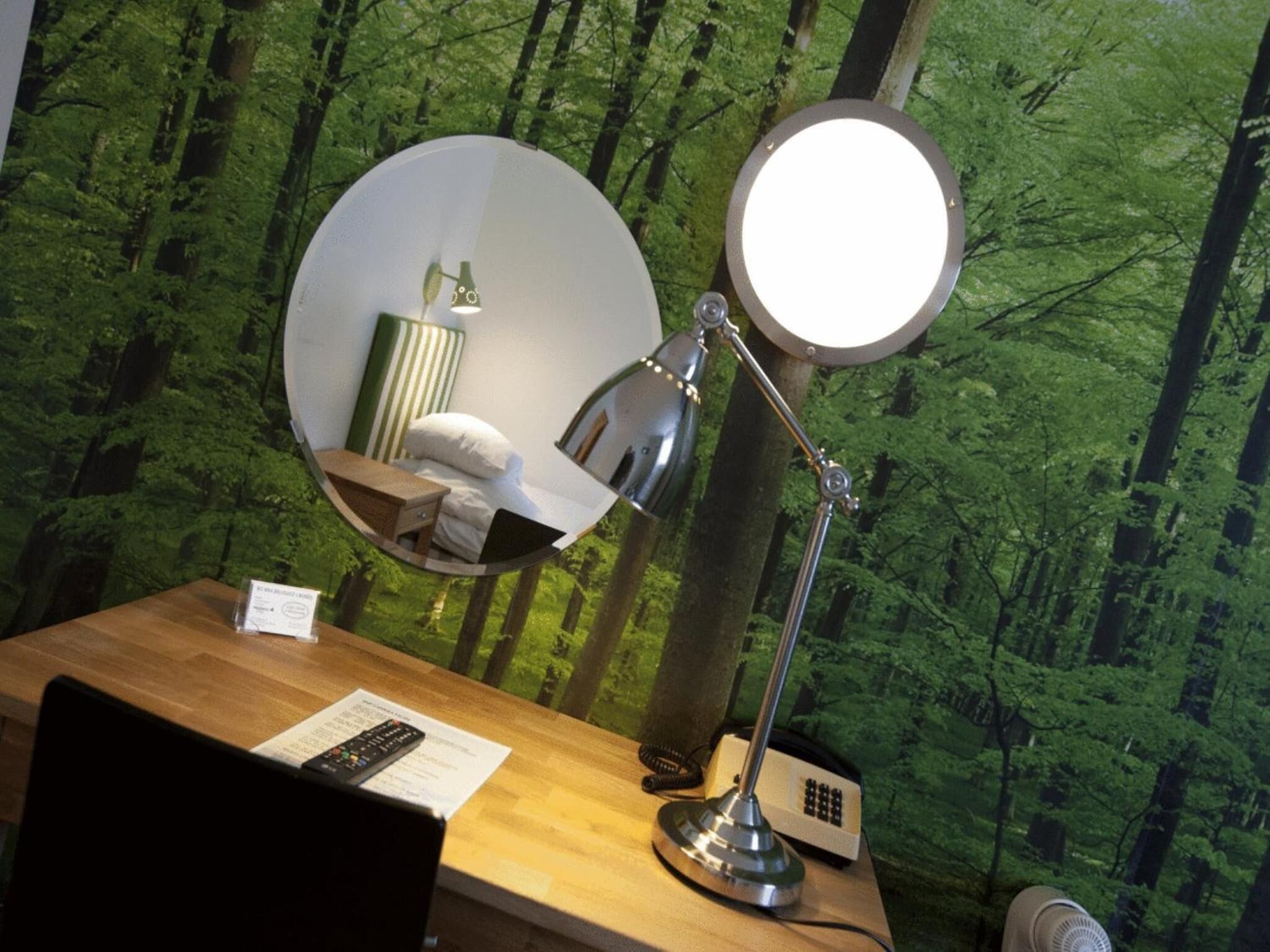 A desk with a mirror, a lamp, a telephone, and a remote control.