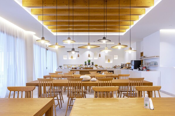 Un comedor moderno con mesas y sillas de madera, iluminación colgante y una cocina al fondo.