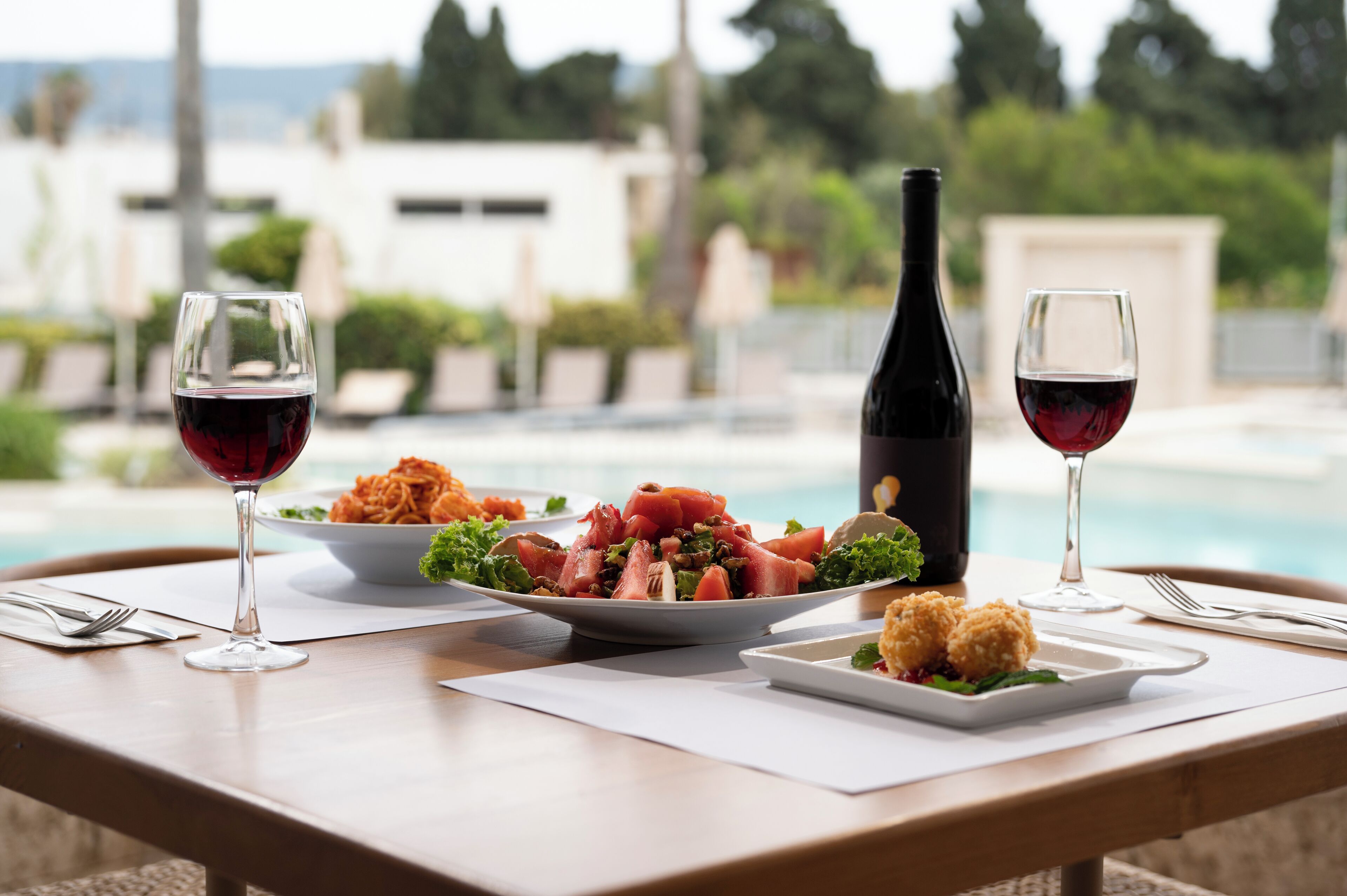 A dining table with plates of food, wine glasses, and a bottle of wine, set against a poolside view.